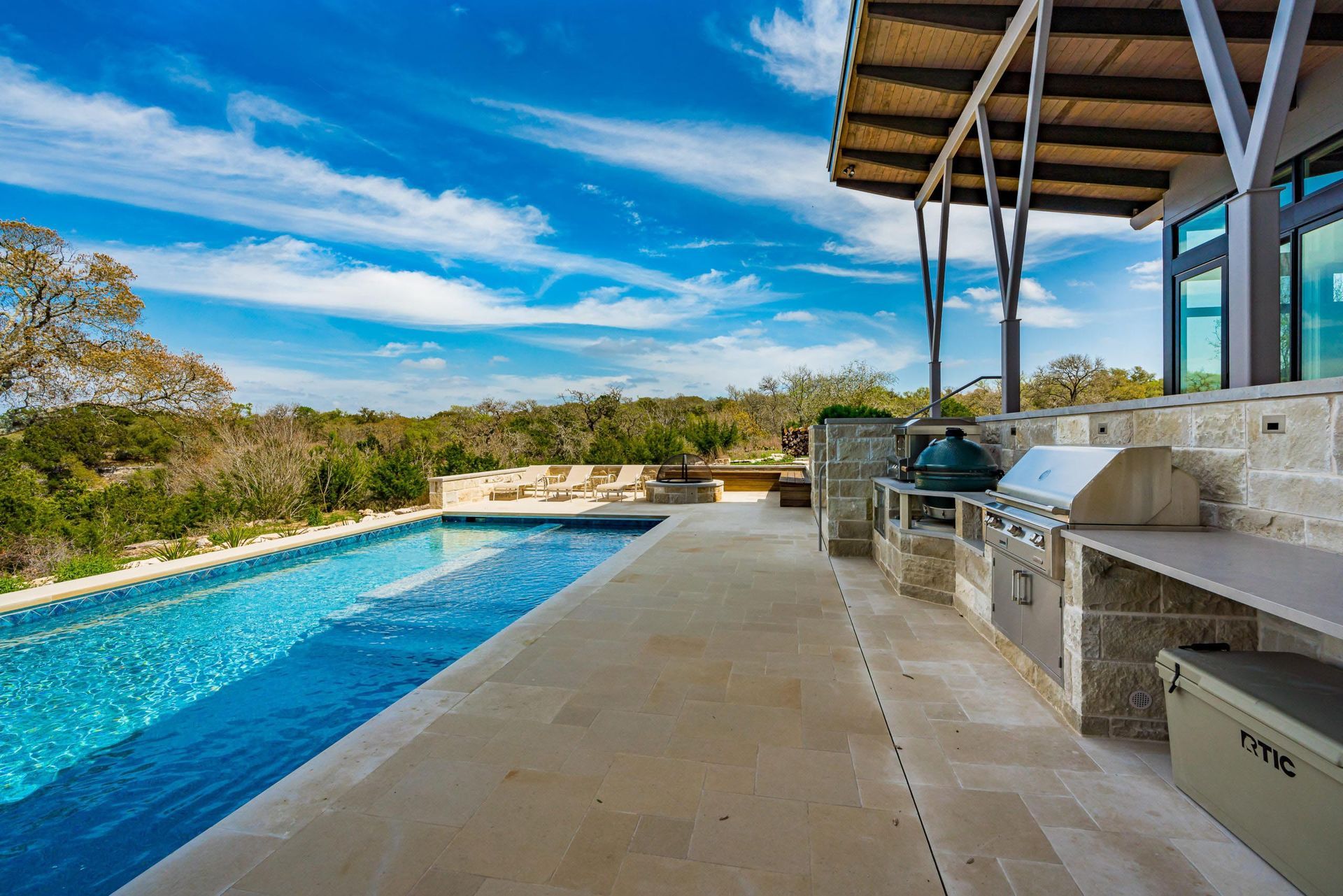 Poolside outdoor kitchen with a blue pool, stone patio, and natural landscape under a bright sky.