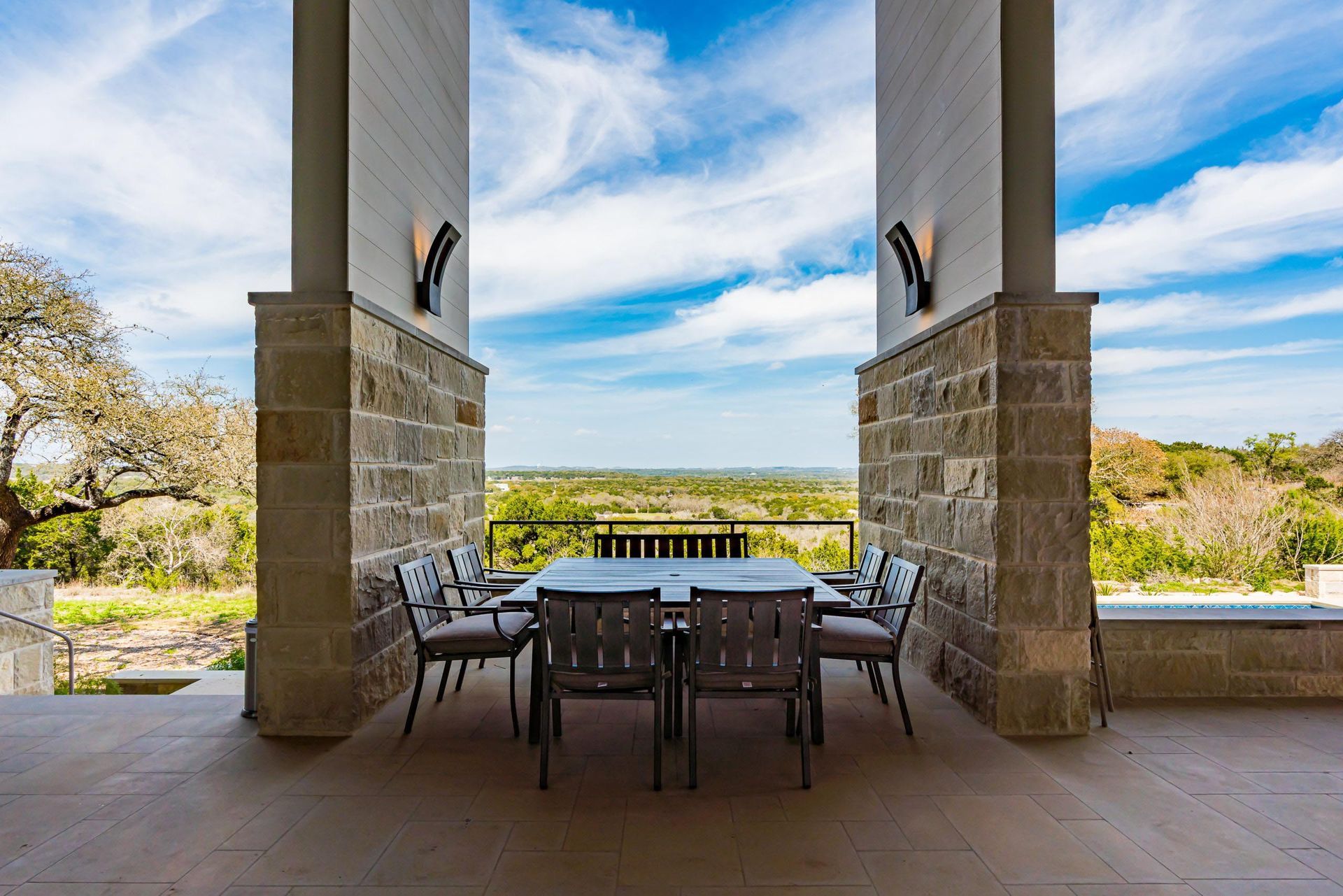 Outdoor dining table and chairs on a covered patio with scenic views, framed by stone columns, under a blue sky.