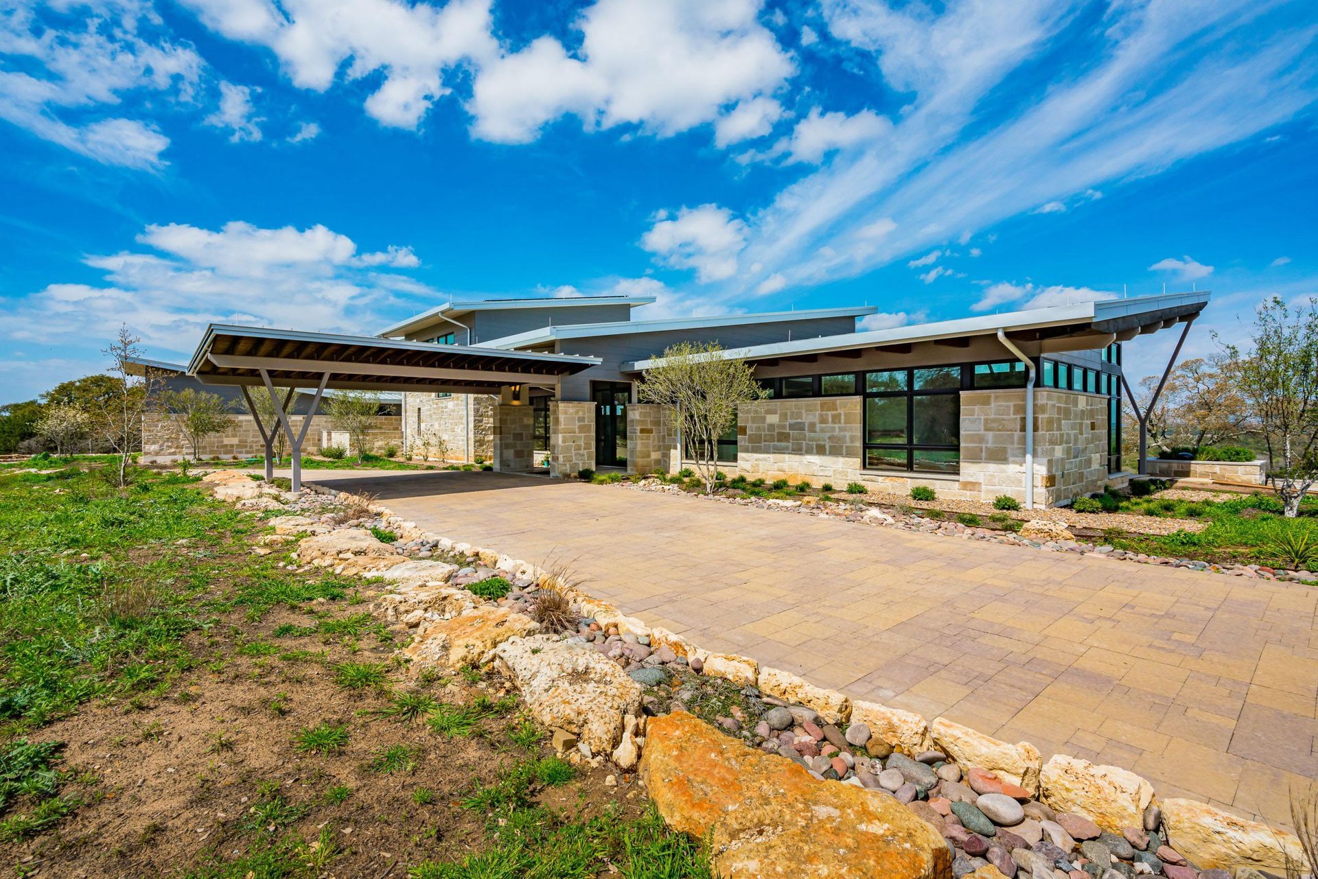 Modern home with angled roof and carport, stone and glass facade, long driveway, blue sky.