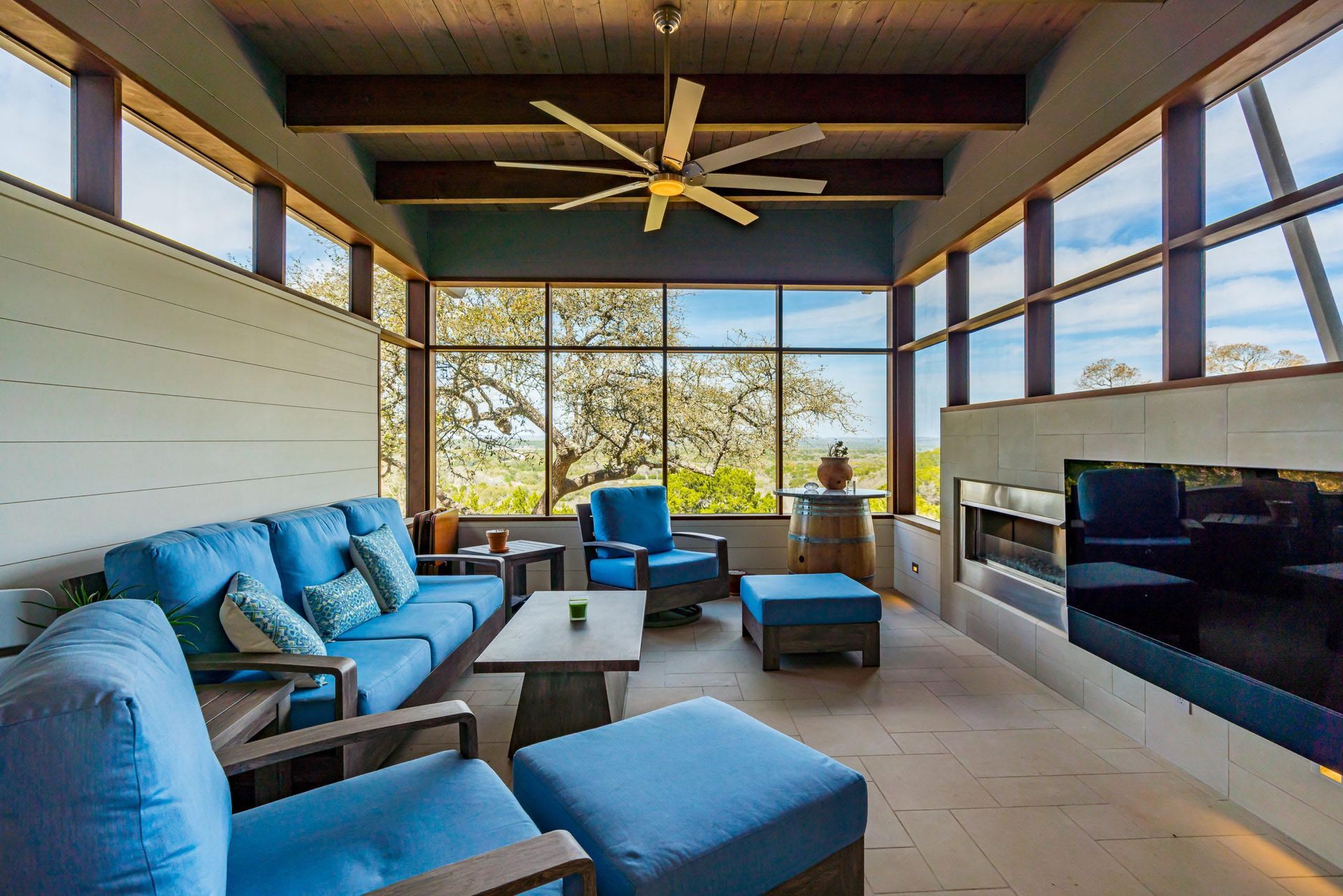 Sunroom with blue outdoor furniture, fireplace, large windows, and a view of trees.