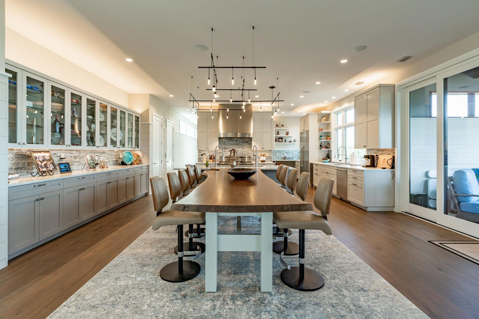 Large kitchen with a long table and chairs on a rug. Cabinets and windows line the walls.