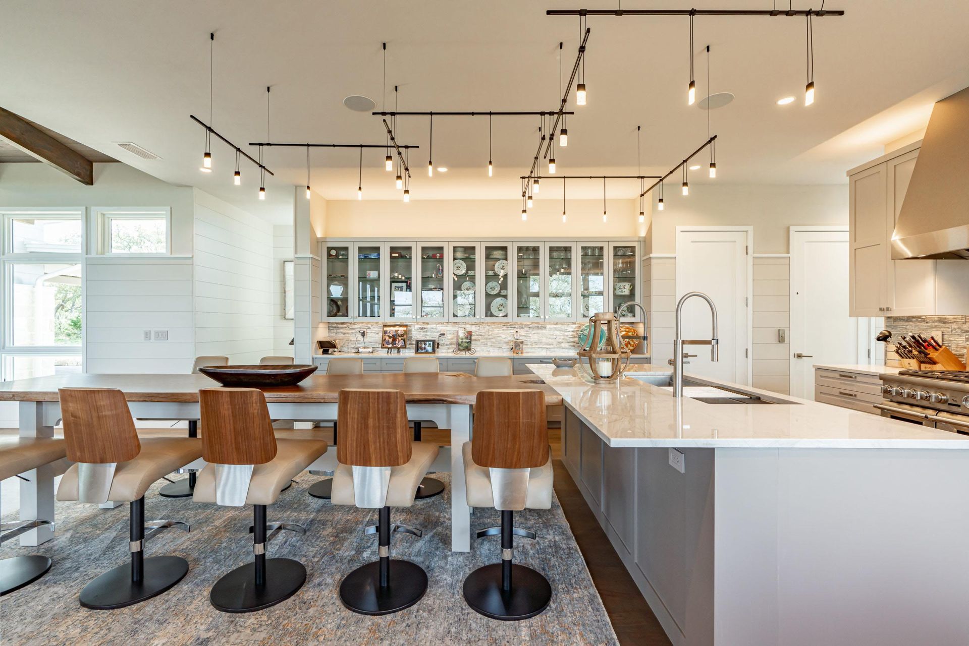 Modern kitchen with long dining table, island, and track lighting. Light cabinets and a stone backsplash.
