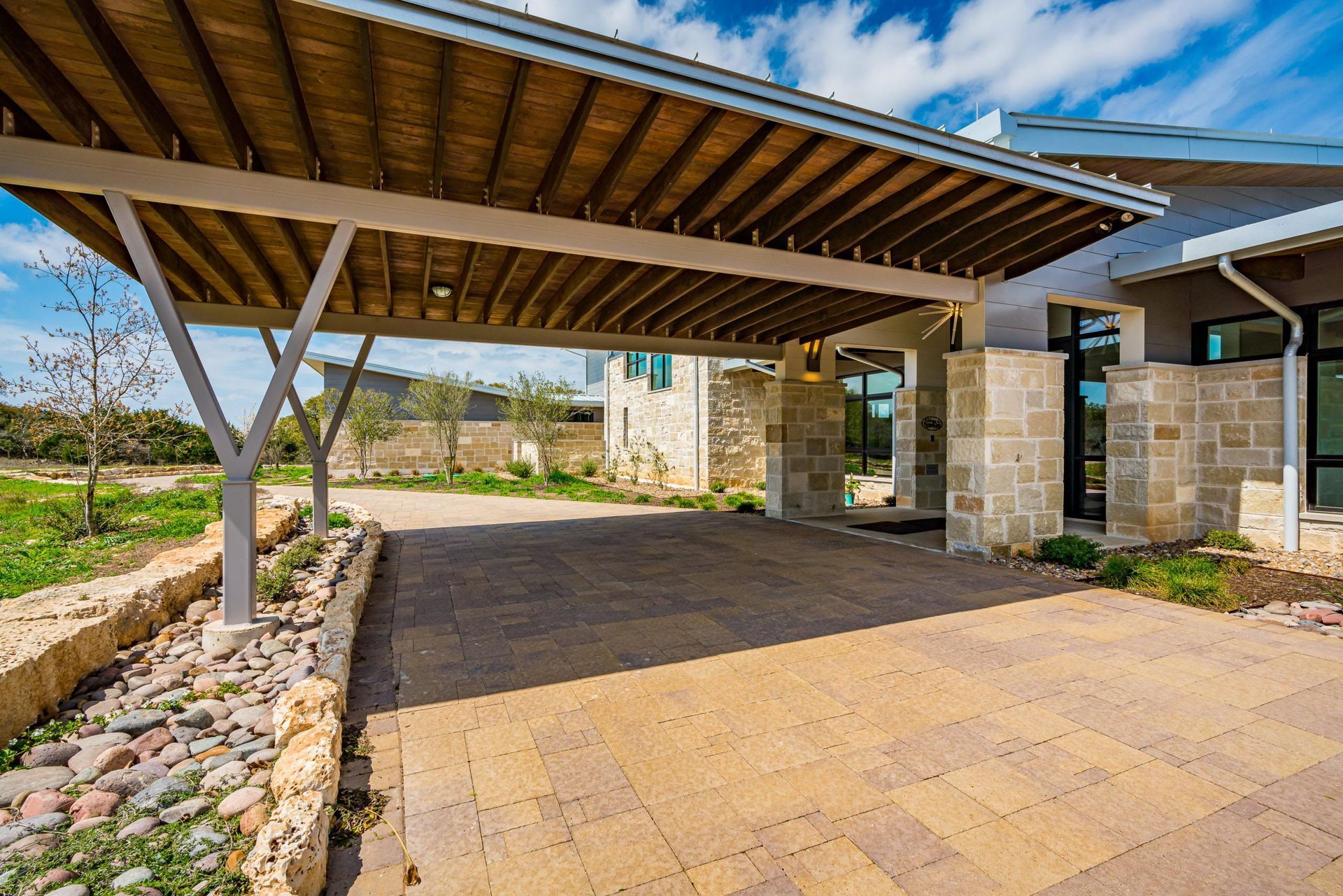 Carport with a wooden ceiling over a paved driveway leading to a stone house.