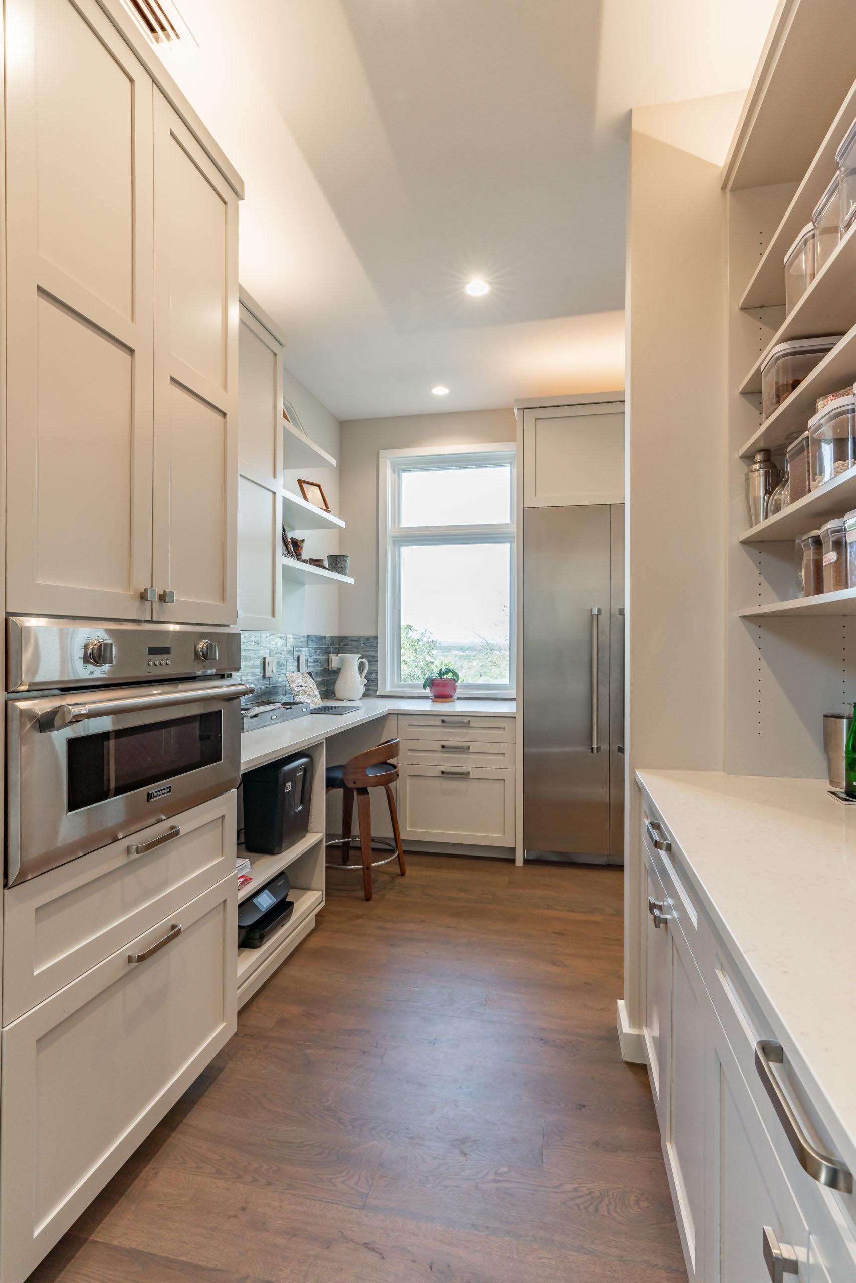 Narrow kitchen with light-colored cabinets, stainless steel appliances, a window, and pantry shelving.