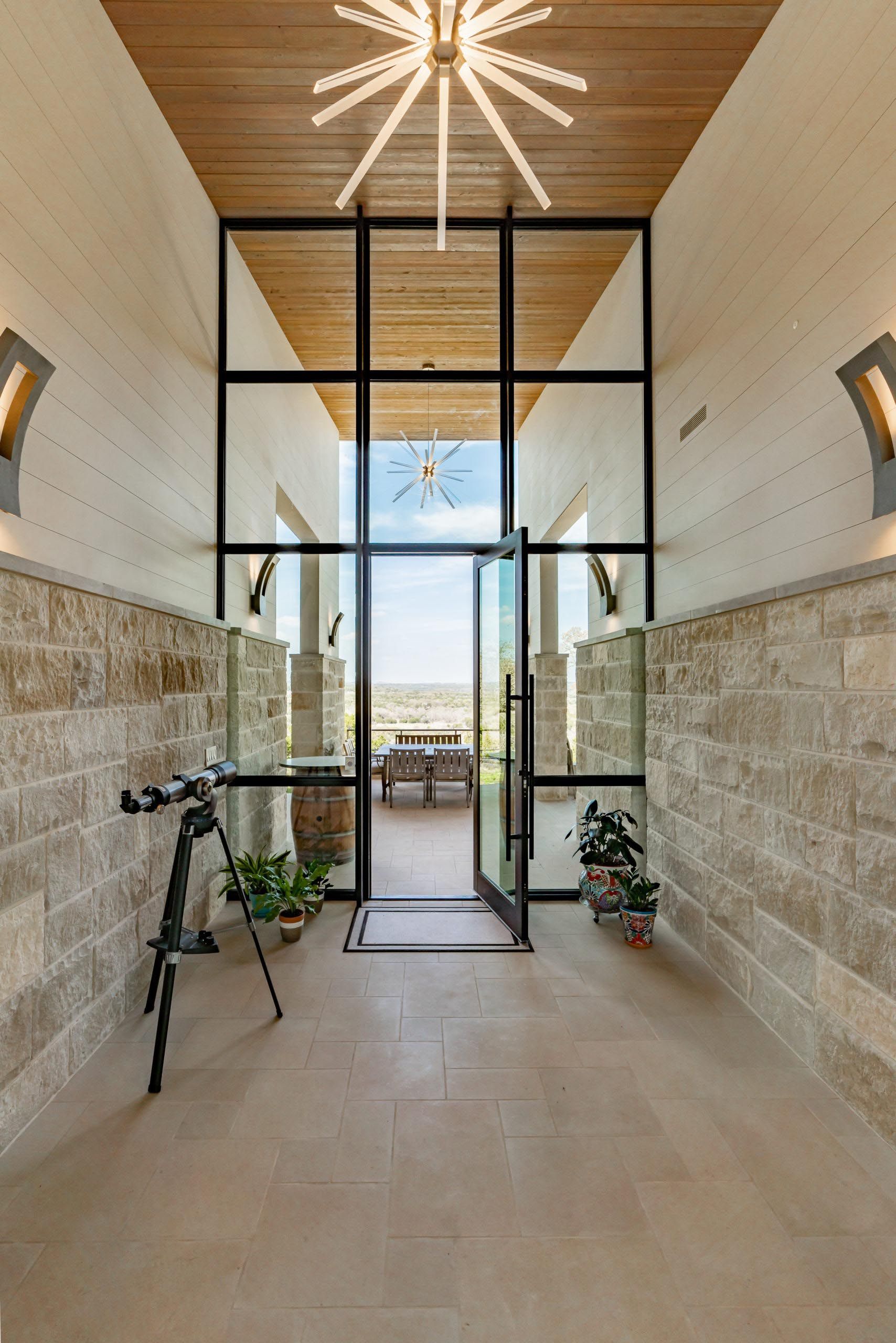 Entryway with glass doors and black frame, light-colored stone walls, wood ceiling, and a view of outdoor patio.