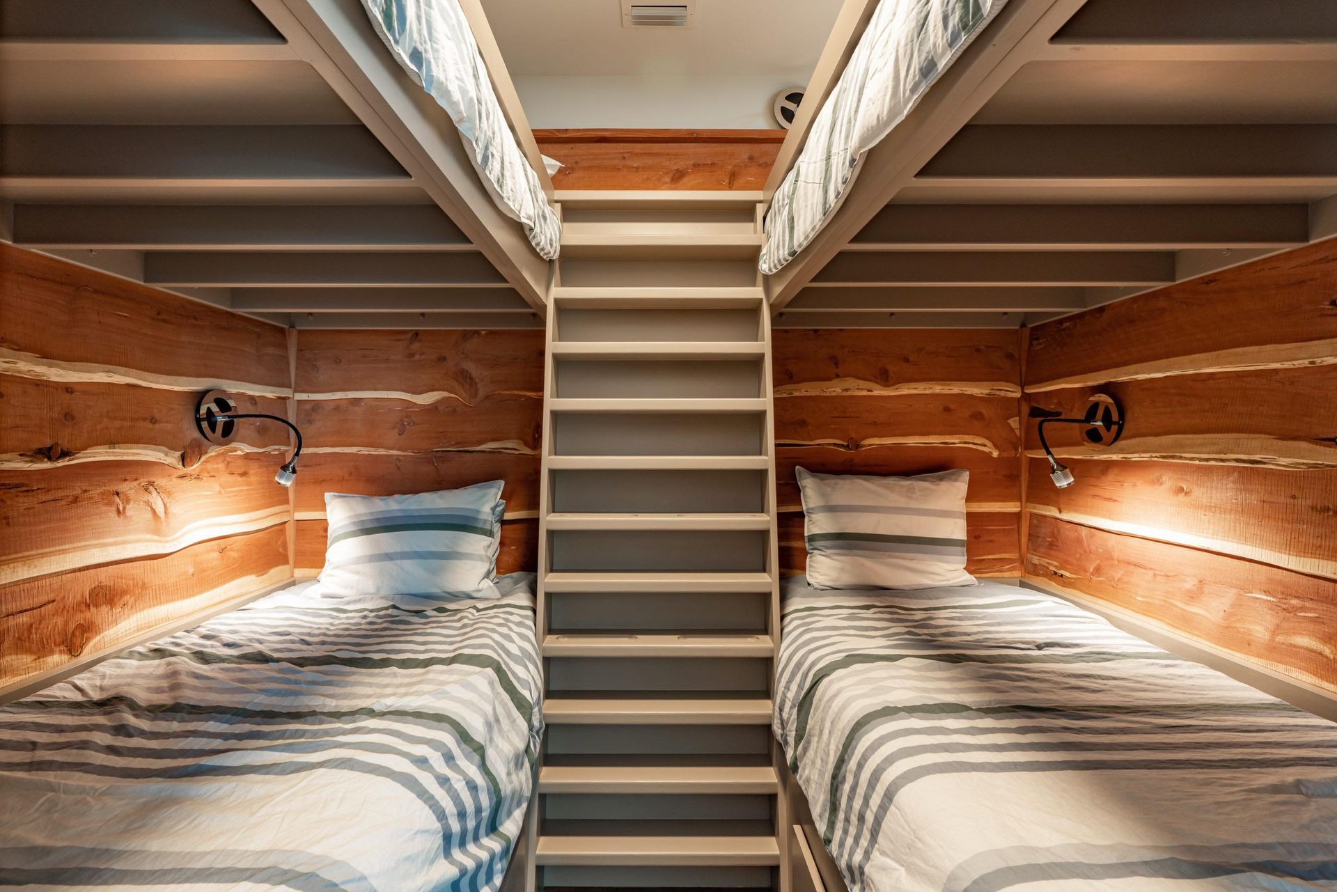 Bunk beds in a cabin-style room. Wooden walls, a ladder, striped bedding, and sconces provide soft light.