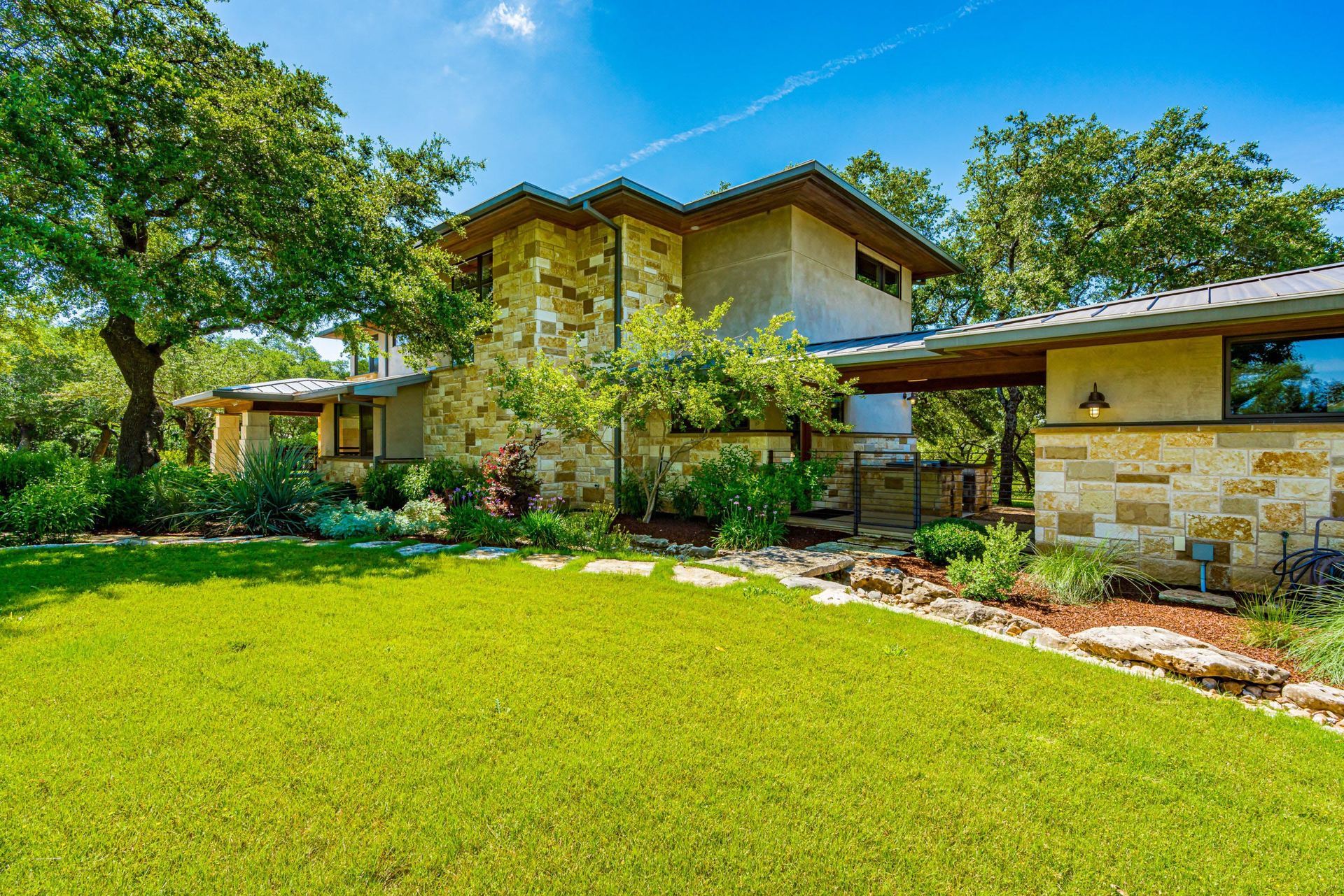 House with stone facade and a green lawn under a blue sky, surrounded by trees.