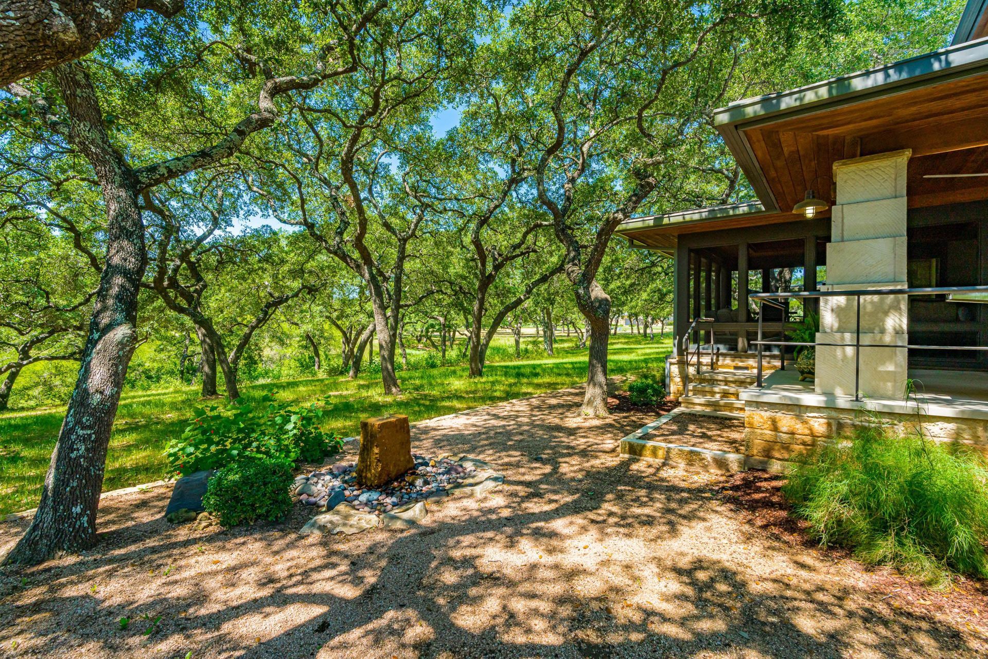 Exterior view of a house with stone columns, porch, and a gravel pathway leading through a green, wooded area.