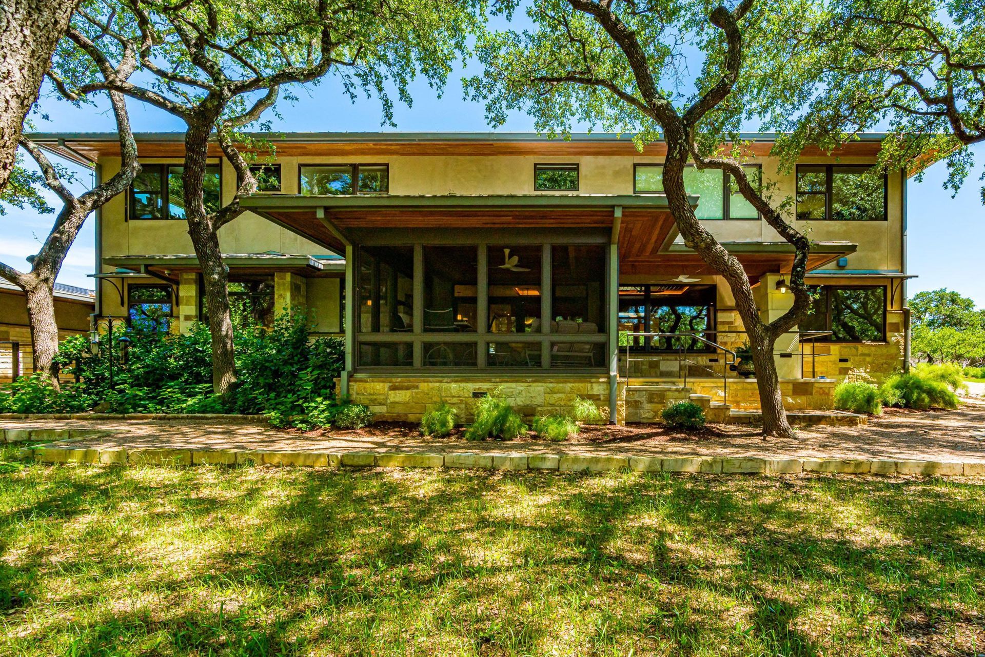 Two-story house with tan siding, large windows, and a shaded porch, framed by trees under a blue sky.