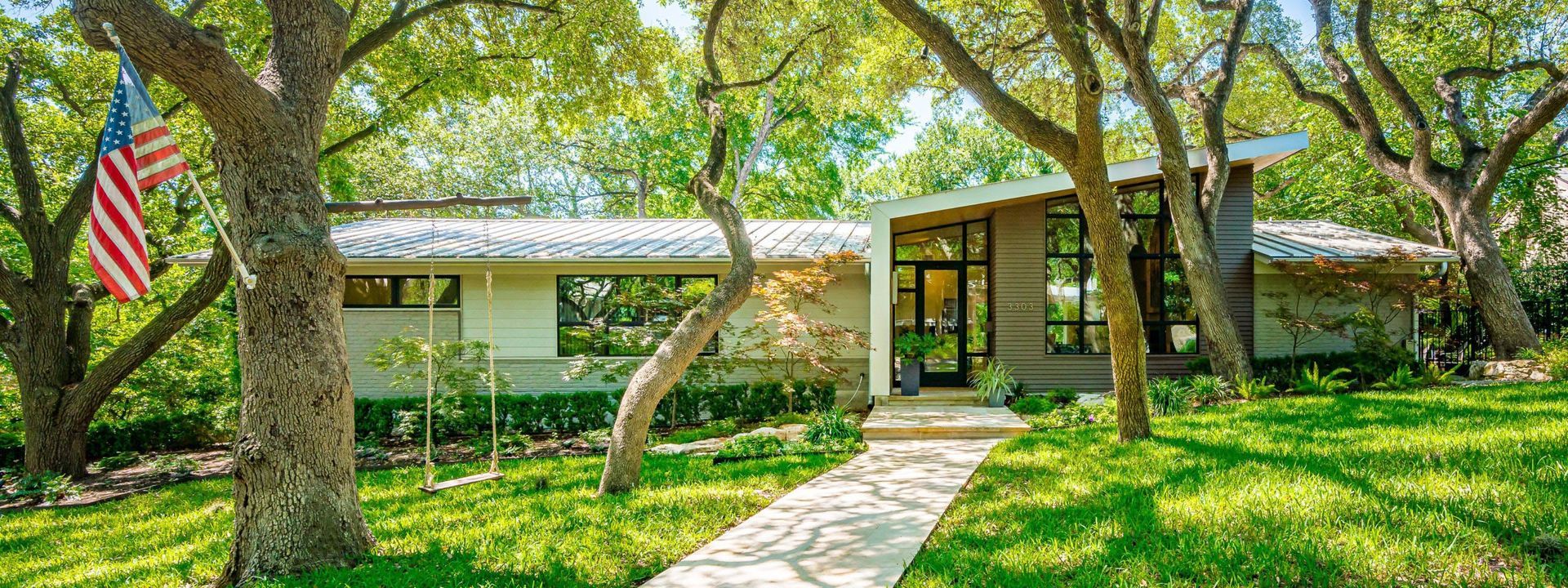 A mid-century modern home with a walkway, surrounded by trees and a waving American flag.