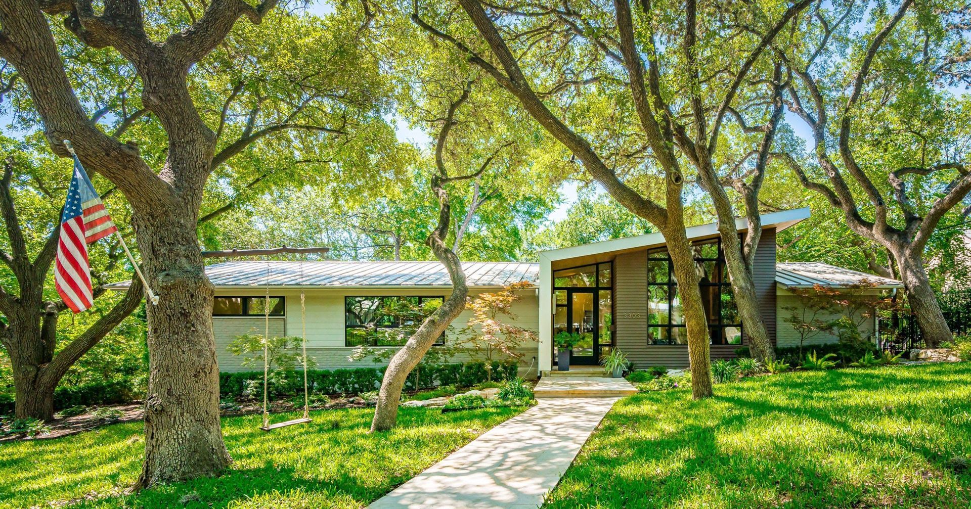 Mid-century modern home with stone accents, nestled amongst trees, American flag, sunny day.