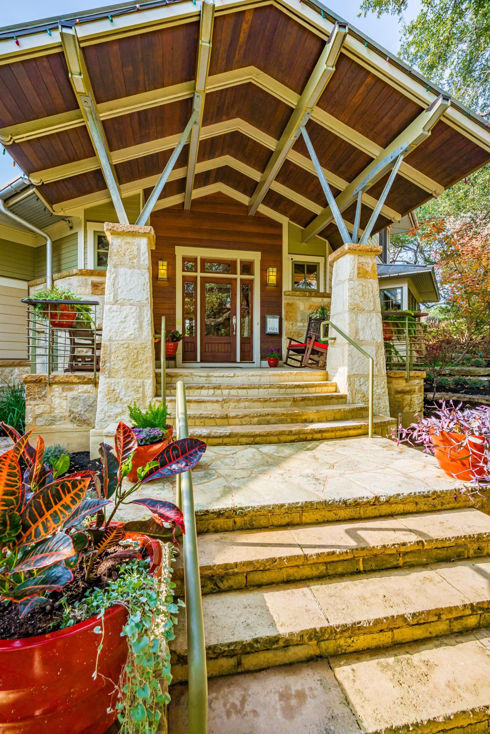 Stone-columned entryway with steps leading to a wooden door under a sloped, wood-paneled awning; potted plants in foreground.