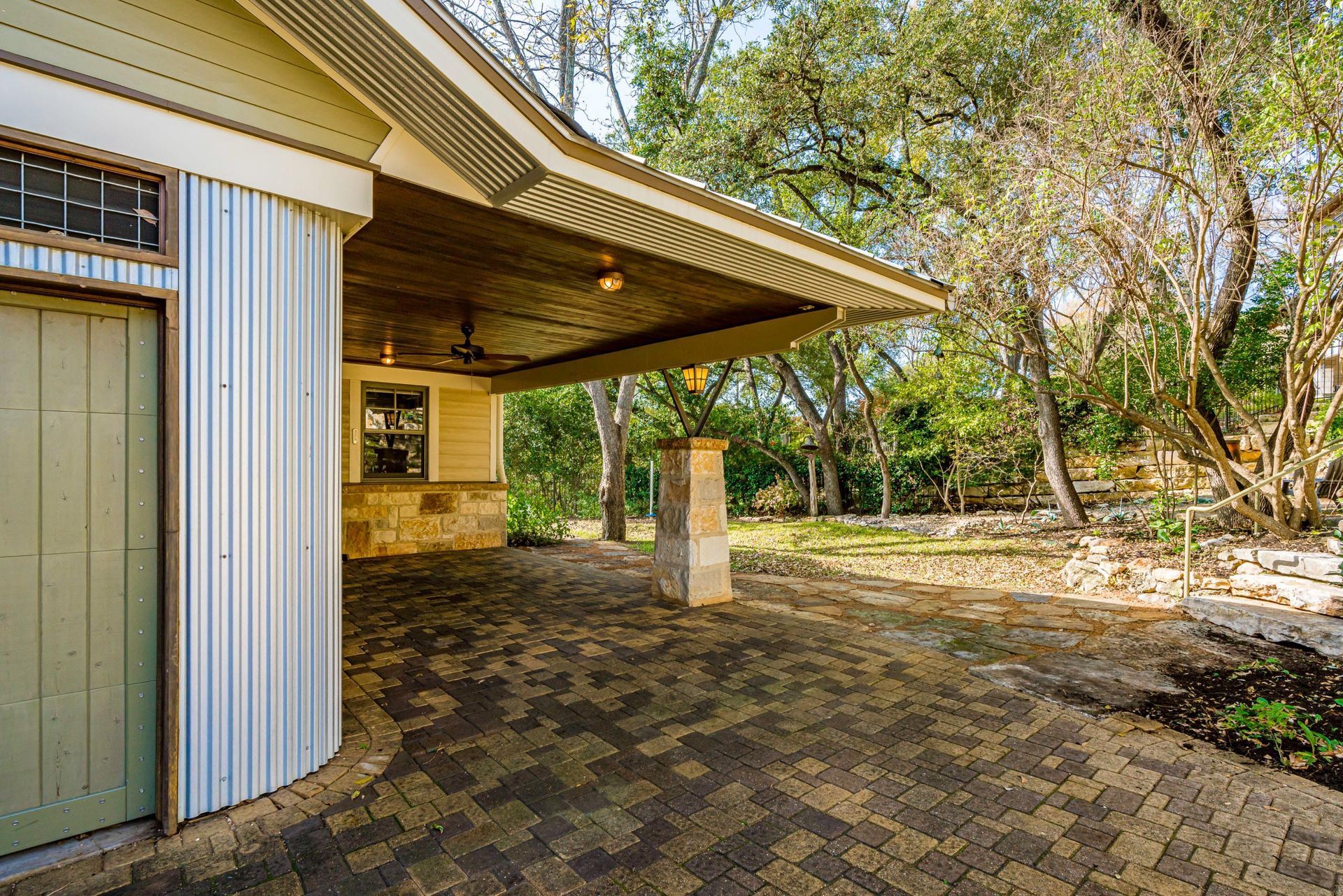 Covered outdoor patio with brick pavers, building with corrugated metal siding and green door, trees in background.