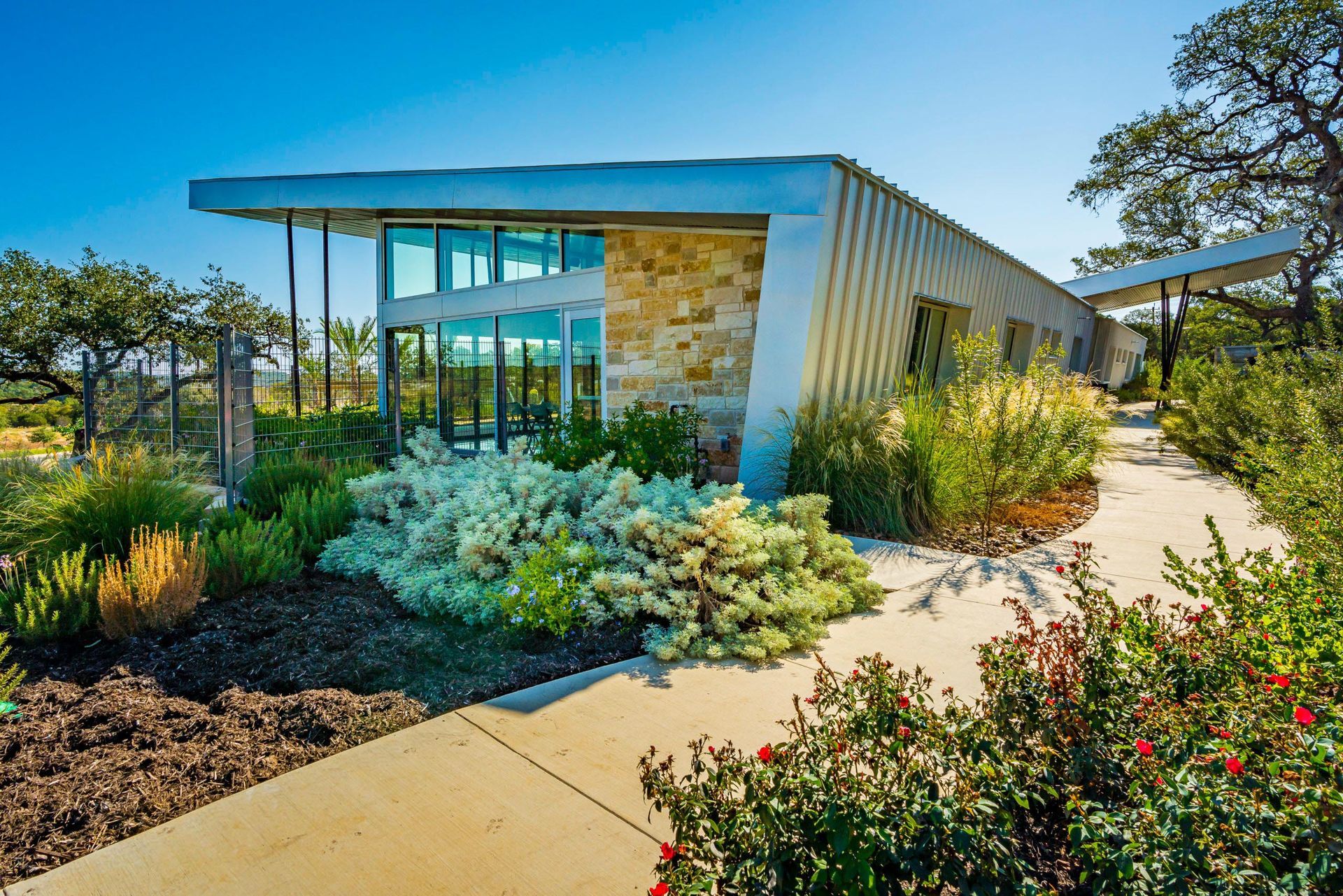 Modern building with glass windows, stone walls, and a metal roof surrounded by greenery under a blue sky.