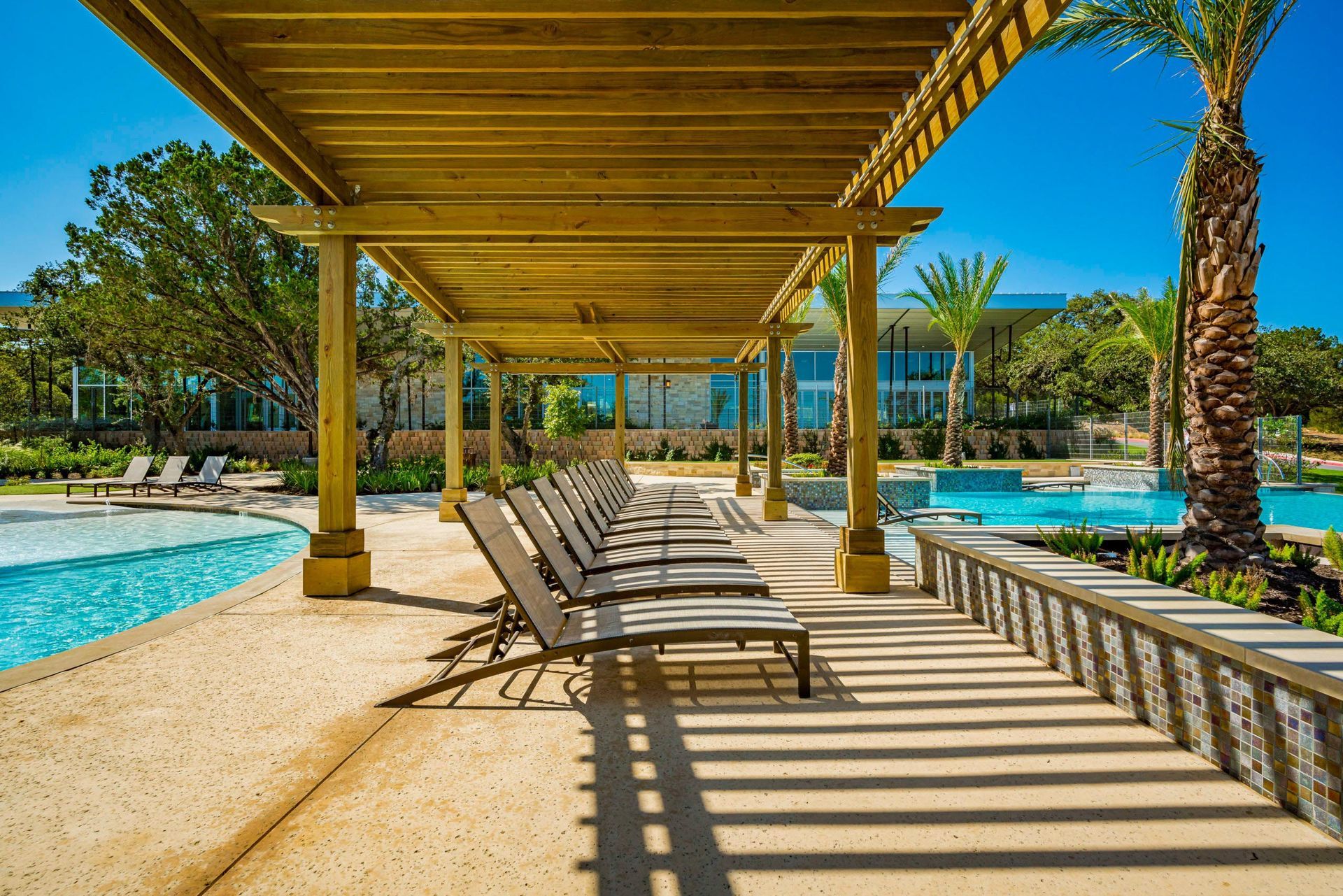 Row of lounge chairs under a wooden pergola next to a pool on a sunny day.
