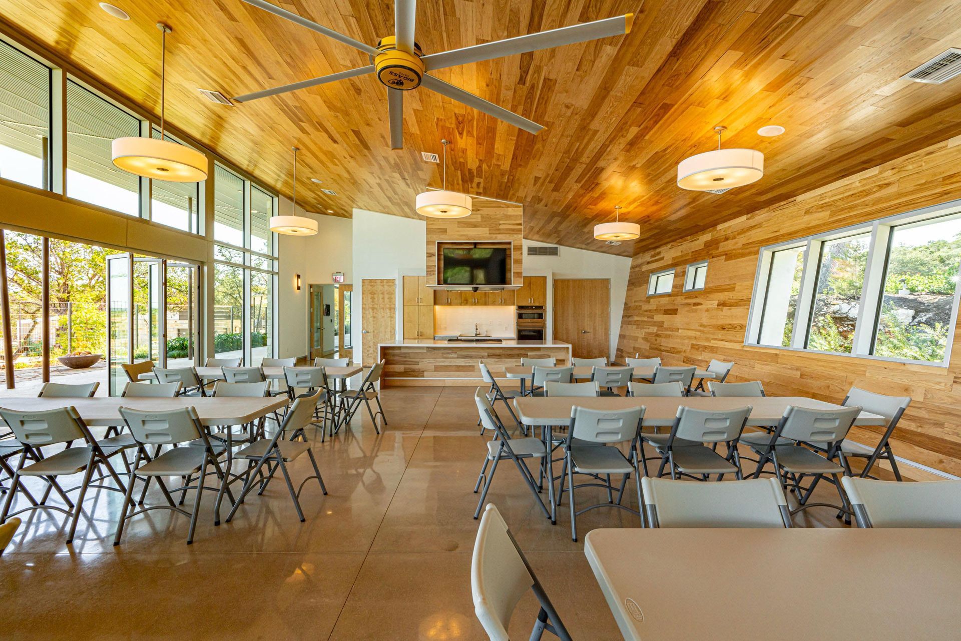 Bright, open community room with tables and chairs, wood ceiling and walls, large windows, and a built-in counter.