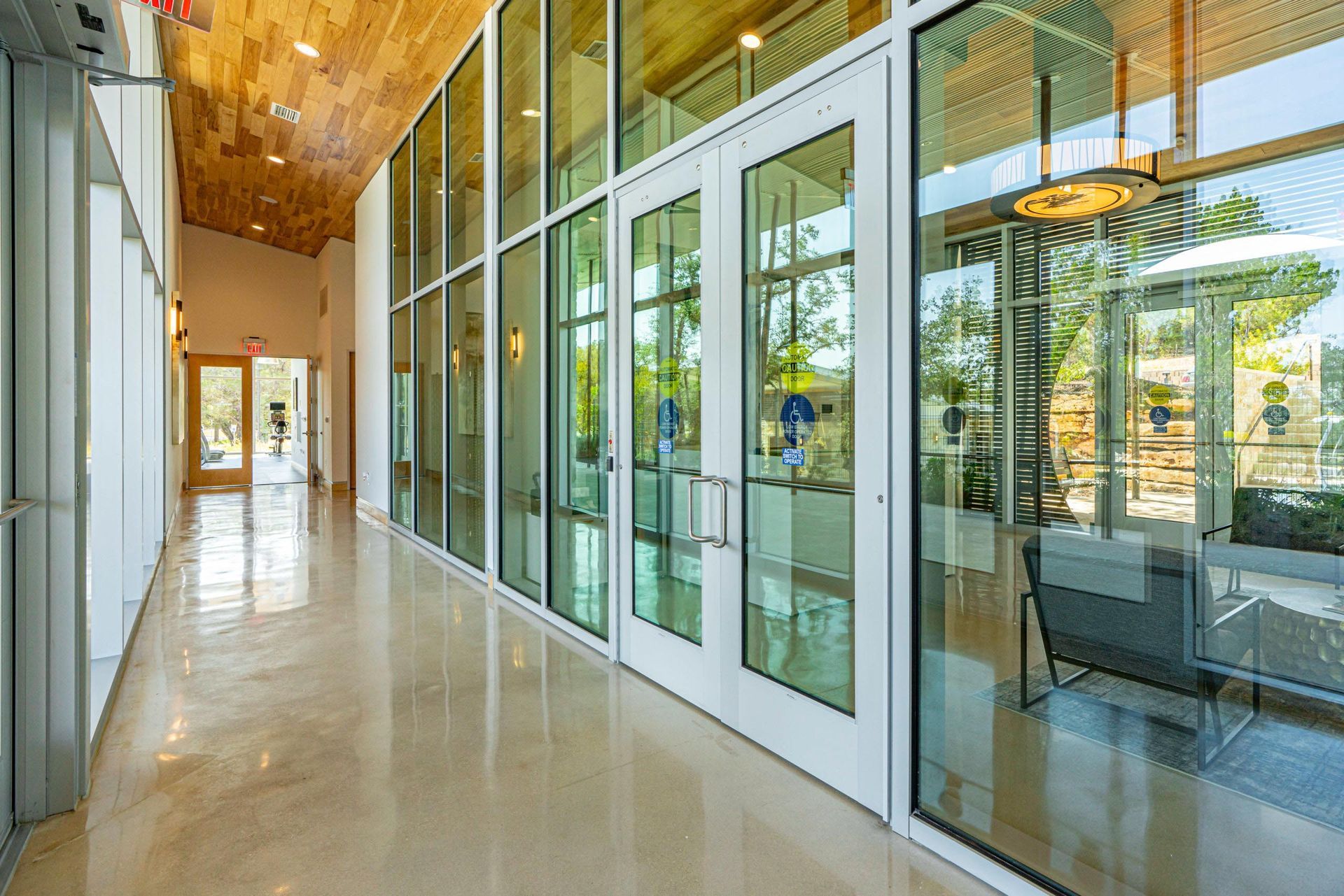 Hallway with large glass doors and windows, light brown floor, and wooden ceiling.
