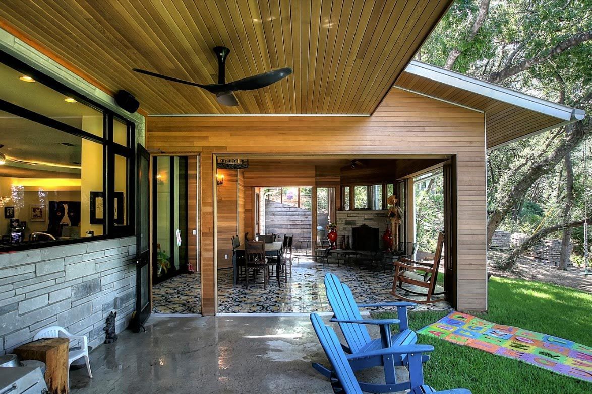Outdoor patio with blue chairs, overlooking a dining area and a room with a fireplace, all under a wooden roof.