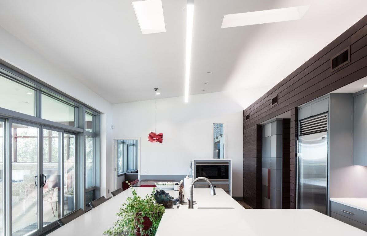 Modern kitchen with large windows, white countertops, and long overhead lighting.