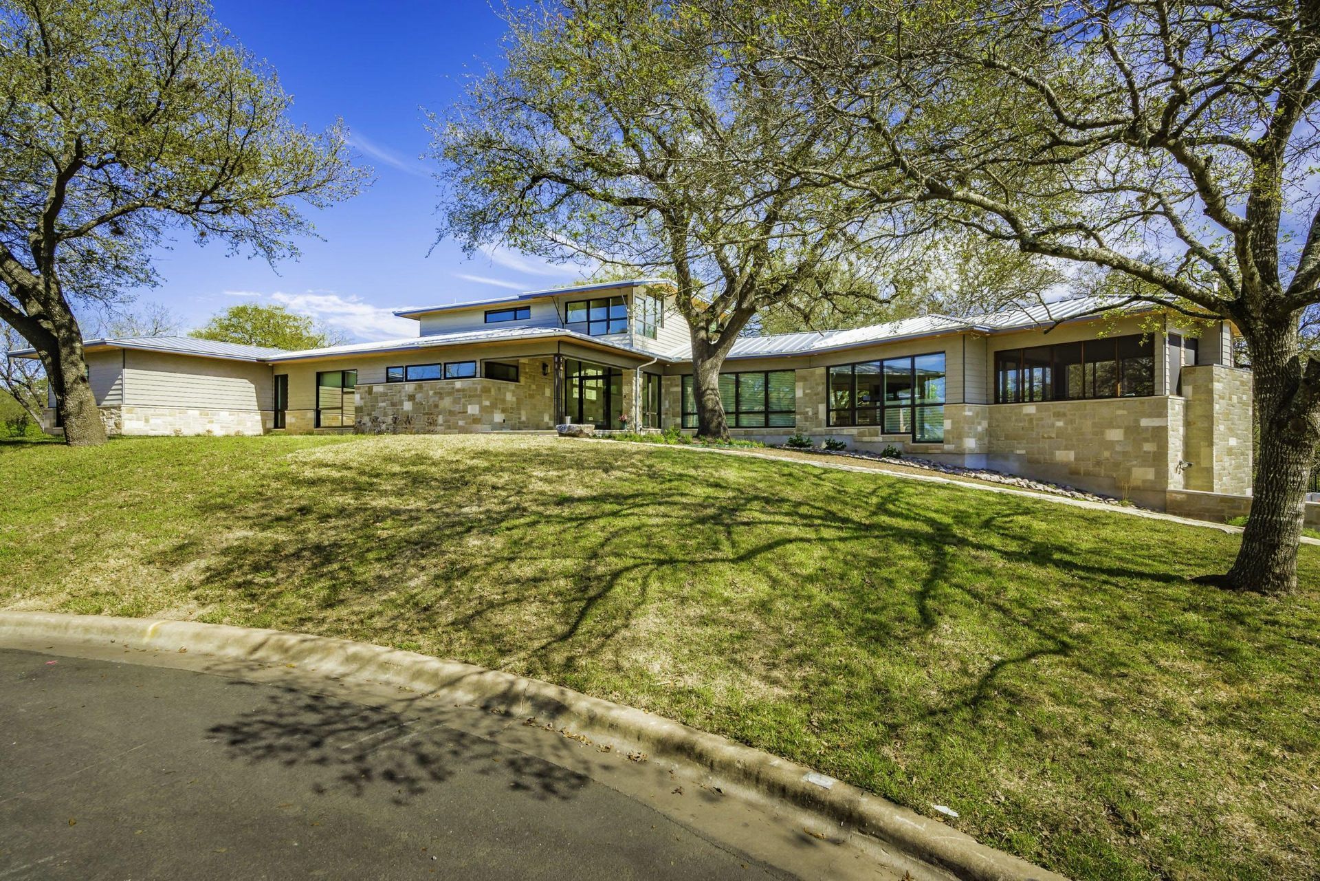 Modern home with stone and glass facade, on a grassy hill, under shade trees.