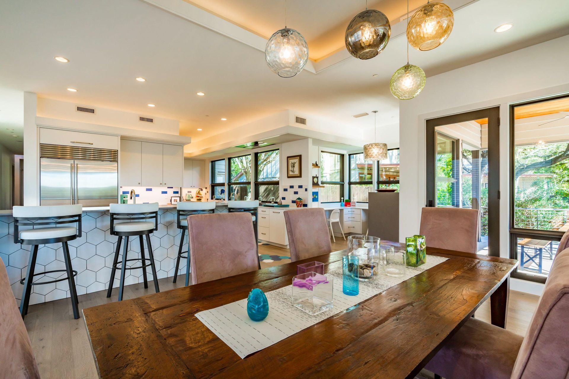 Dining room with wooden table, pink chairs, and modern light fixtures, open to a white kitchen.