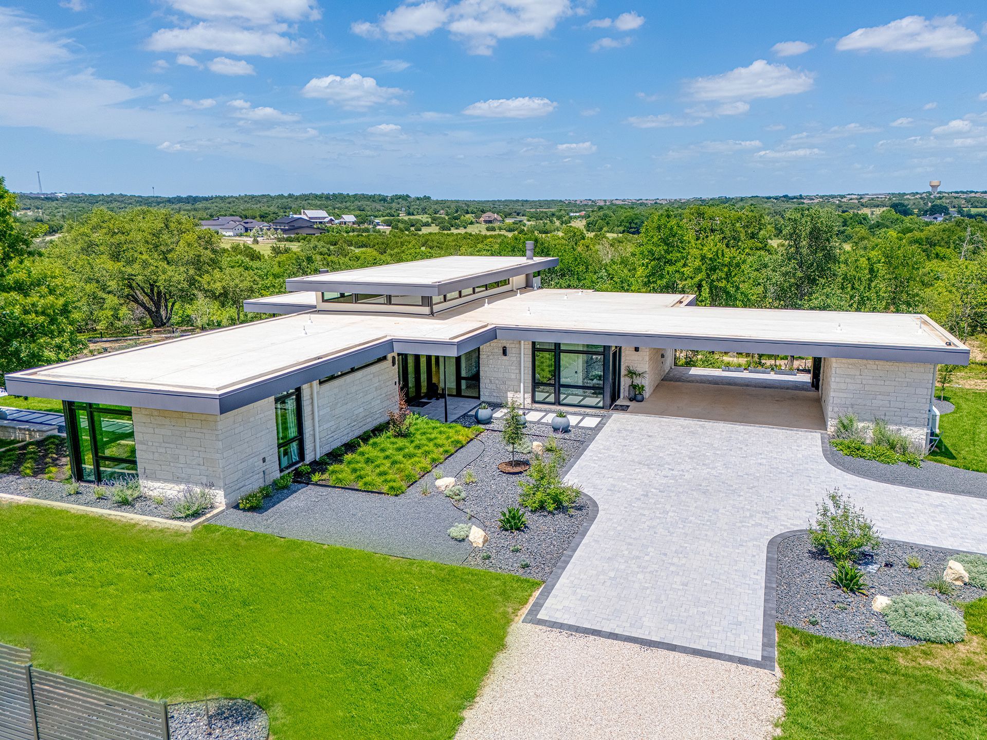 Modern, single-story house with a flat roof and carport, set in a green landscape with a long driveway.