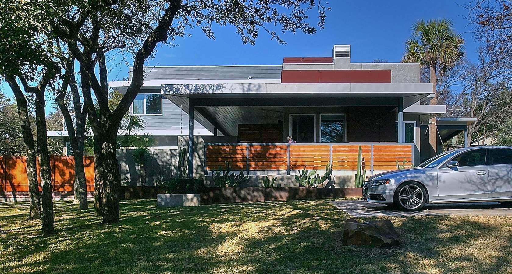 Modern home exterior with a silver car parked on the driveway, a wooden fence, and green trees.