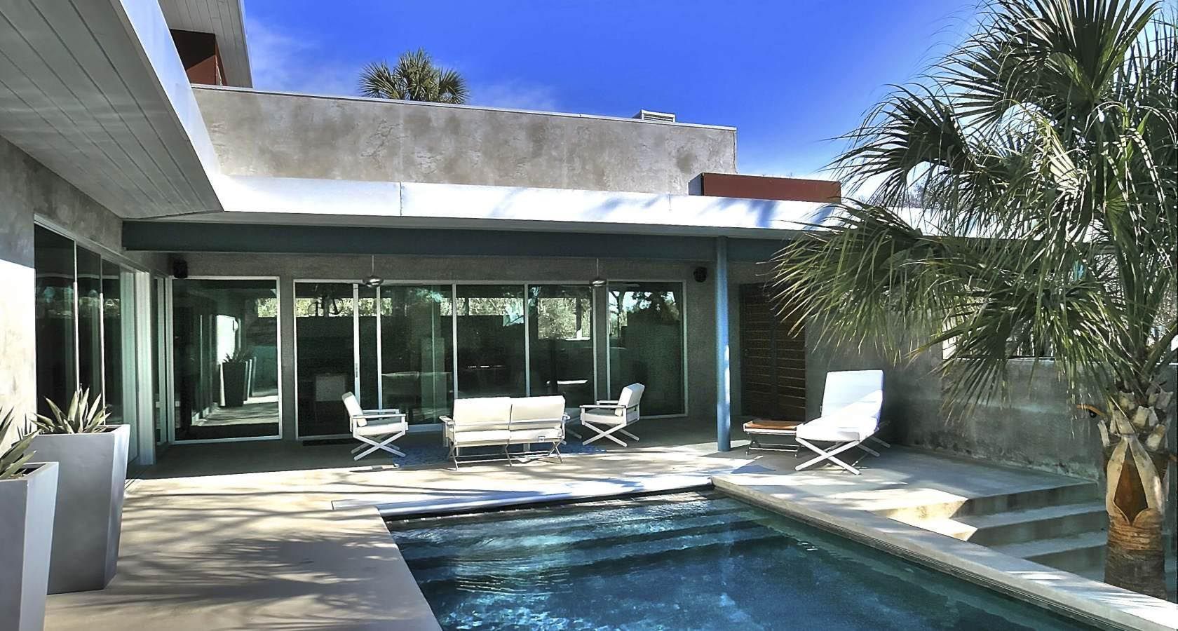 Pool area with modern house and patio furniture. Blue pool, palm tree, and clear sky.