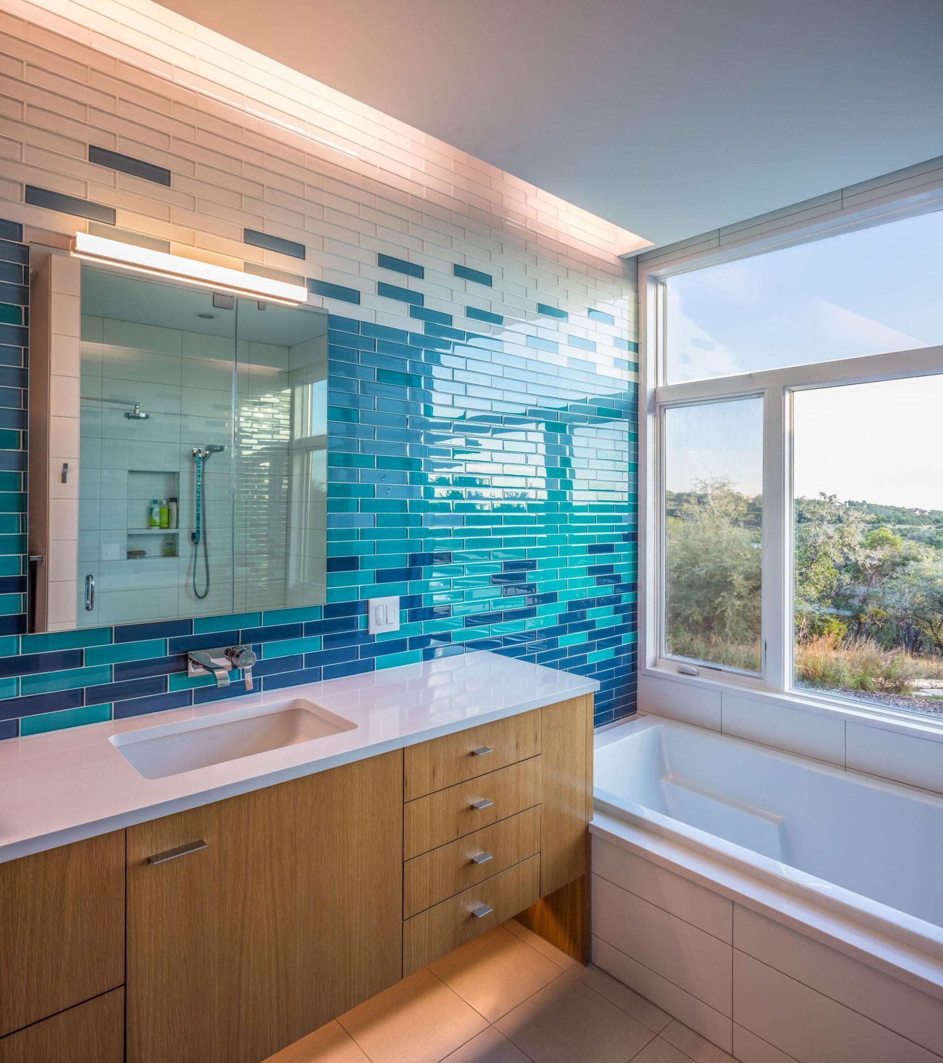 Modern bathroom with blue tile accent wall, wood vanity, large window overlooking landscape.