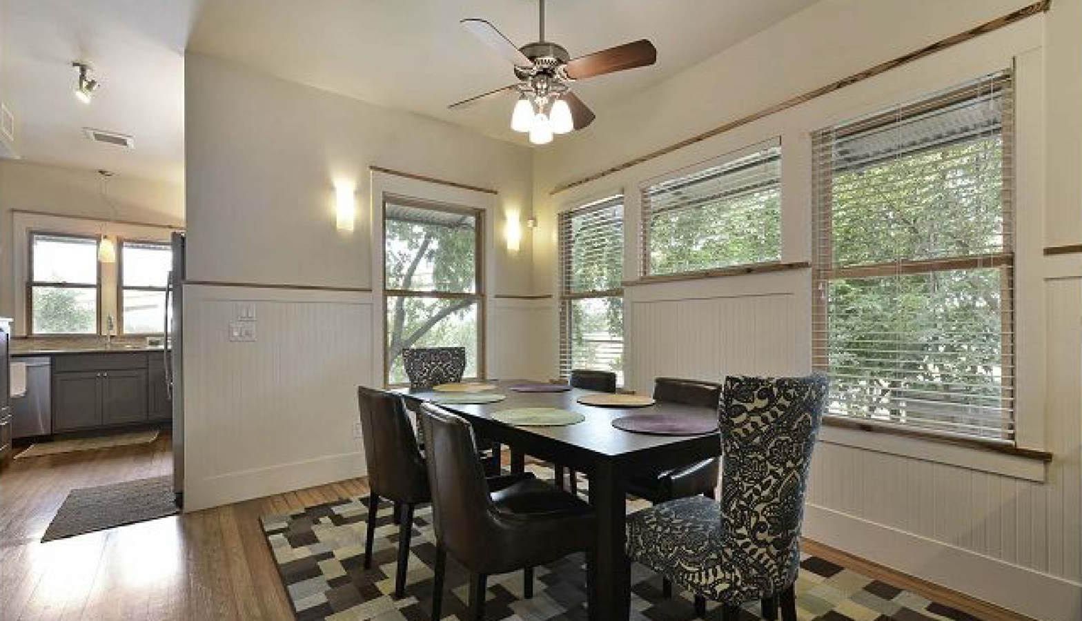 Dining room with a table and chairs, adjacent to a kitchen area. Windows provide outside views.