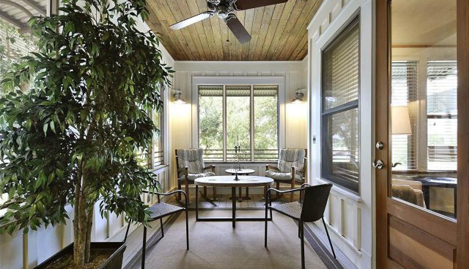 Sunroom with wood ceiling, furniture, large potted tree, and windows overlooking greenery.