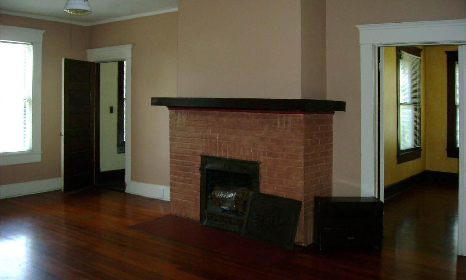 Empty living room with brick fireplace, dark wood floor, and doorway to another room.