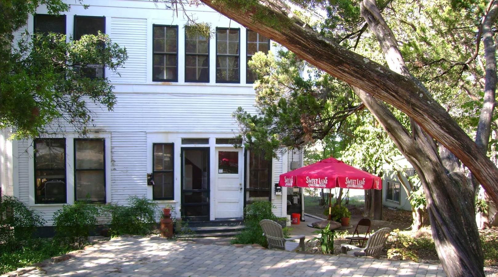 White two-story building with a front porch under trees and a red umbrella.