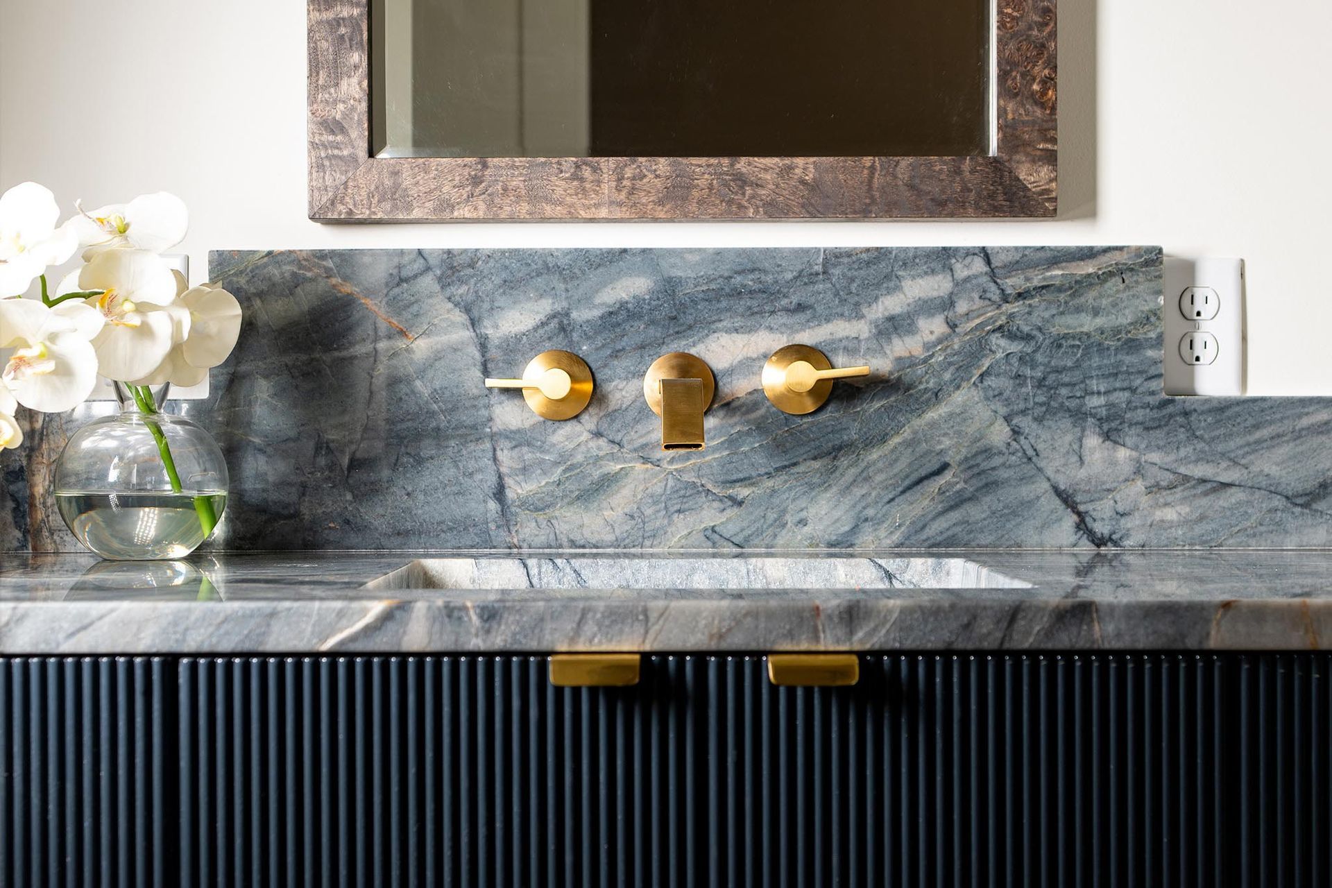 Bathroom vanity with blue veined marble countertop, gold fixtures, and a glass vase with white flowers.