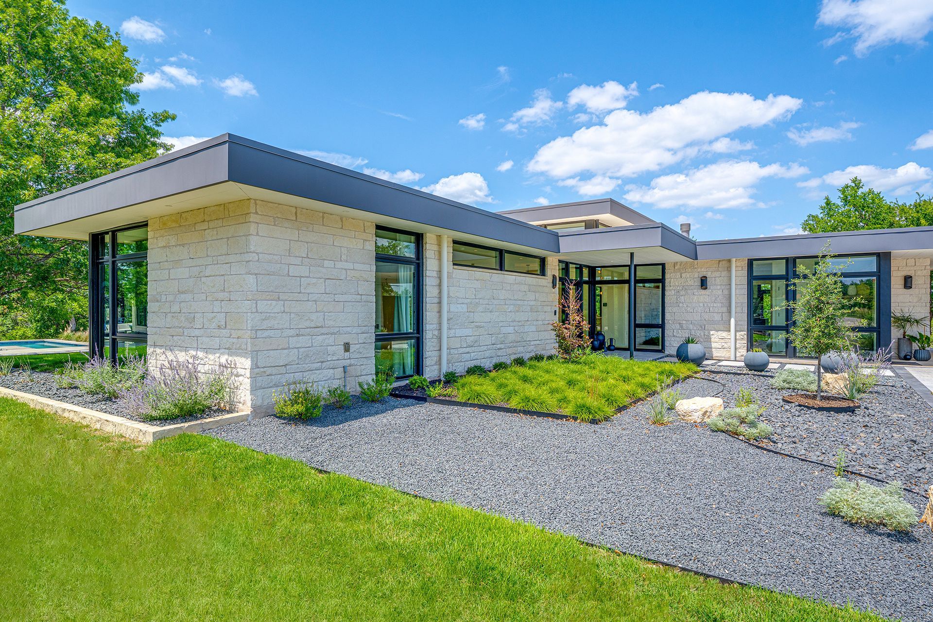 Modern single-story home with a flat roof, large windows, and a black gravel and plant landscaping on a sunny day.