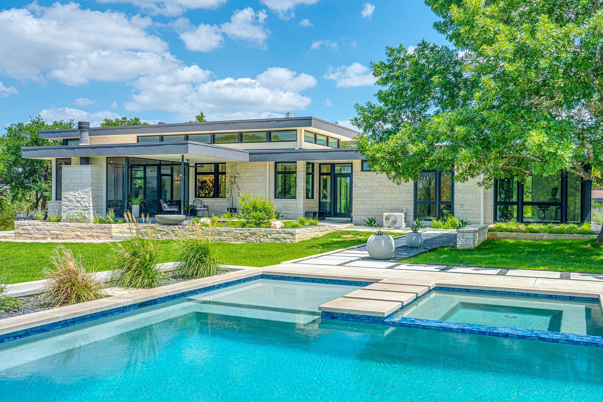 Modern home with pool; light stone exterior, green lawn, large windows, blue sky.