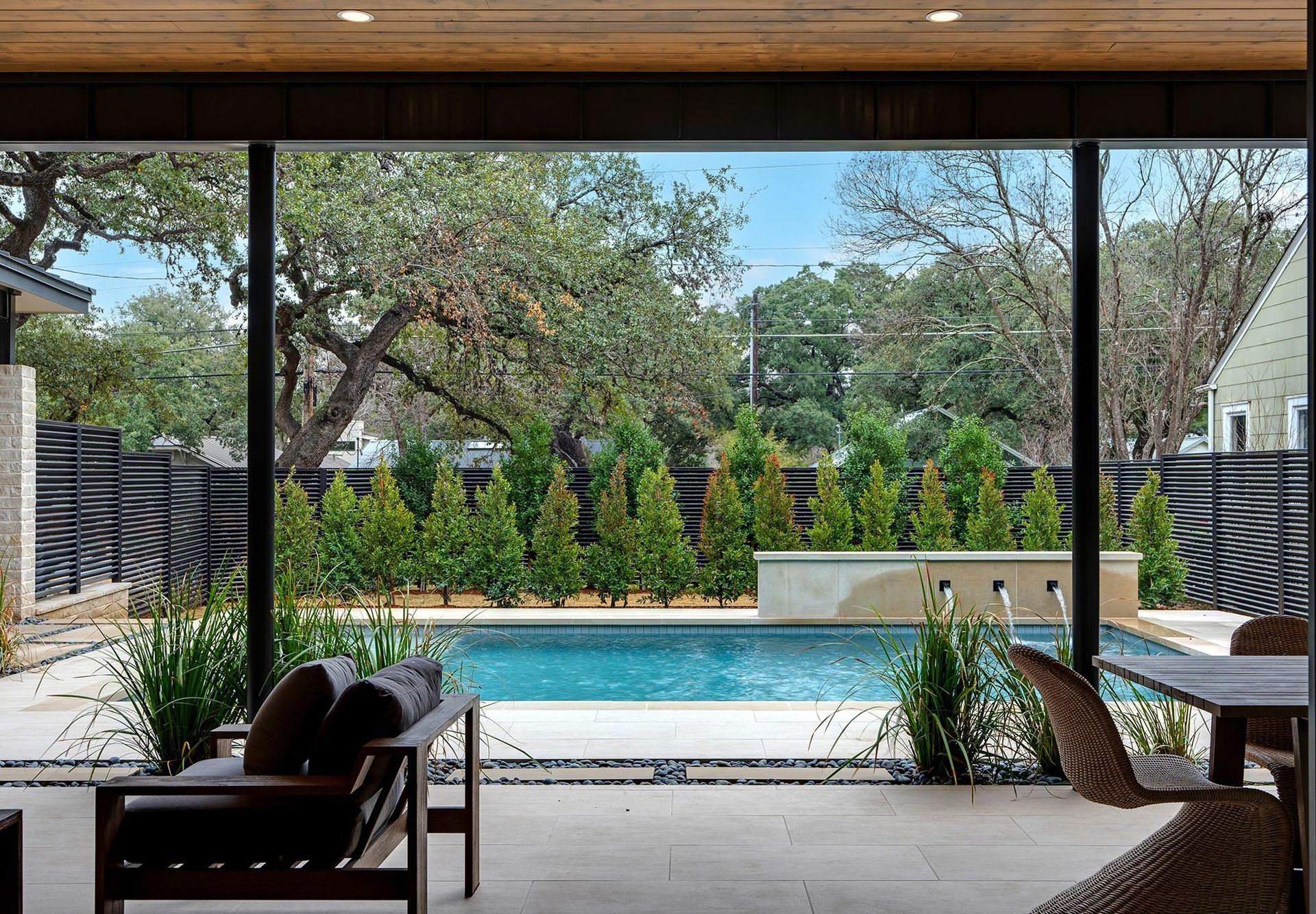Patio overlooking a swimming pool and lush landscaping. Black-framed windows, wood ceiling.