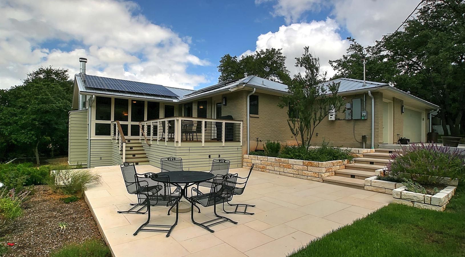 House with patio, dining set, and landscaping under a blue sky with fluffy clouds. Solar panels on roof.