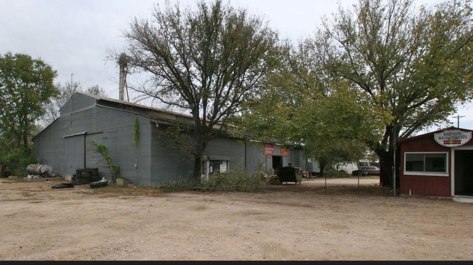 Gray corrugated metal building with chimney, trees, and small red shop on a cloudy day.