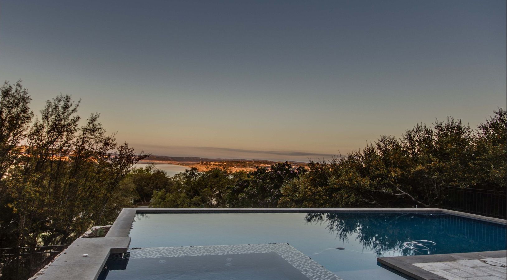 Infinity pool overlooking a town and bay at dusk, surrounded by trees.