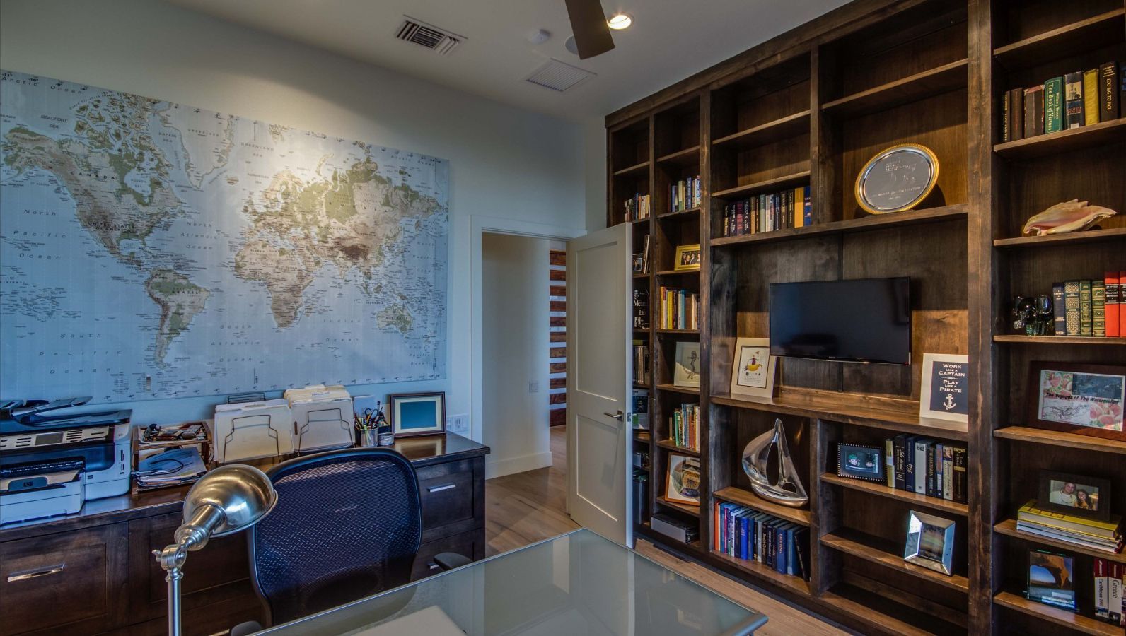 Office with built-in bookshelves, world map, desk, and printer. Doorway leads to a hallway.