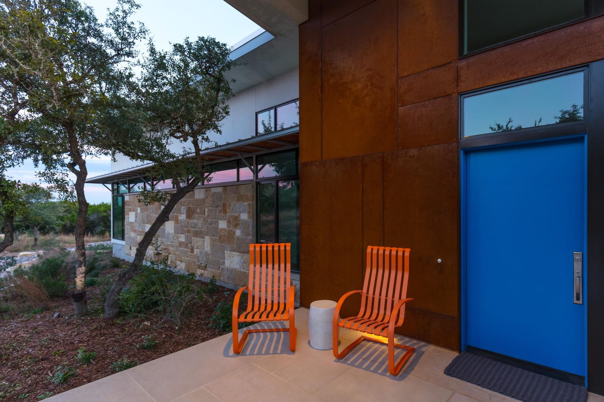 Modern home exterior with orange chairs, blue door, and rust-colored metal siding.