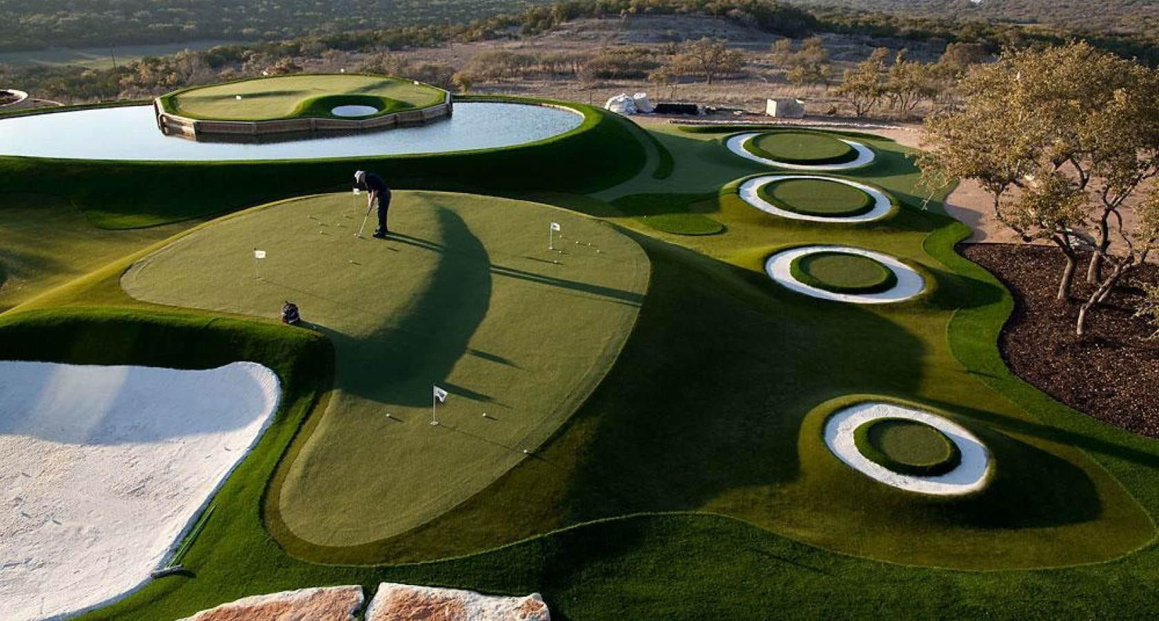 Golfer on a green with various putting greens and water feature at a golf course.