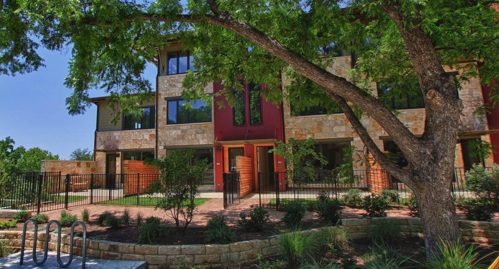 Townhouses with stone facade, under a large tree; blue sky, landscaping.
