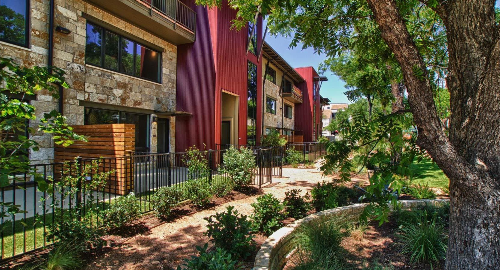 Exterior view of a multi-story apartment building with stone and red accents, surrounded by landscaping.
