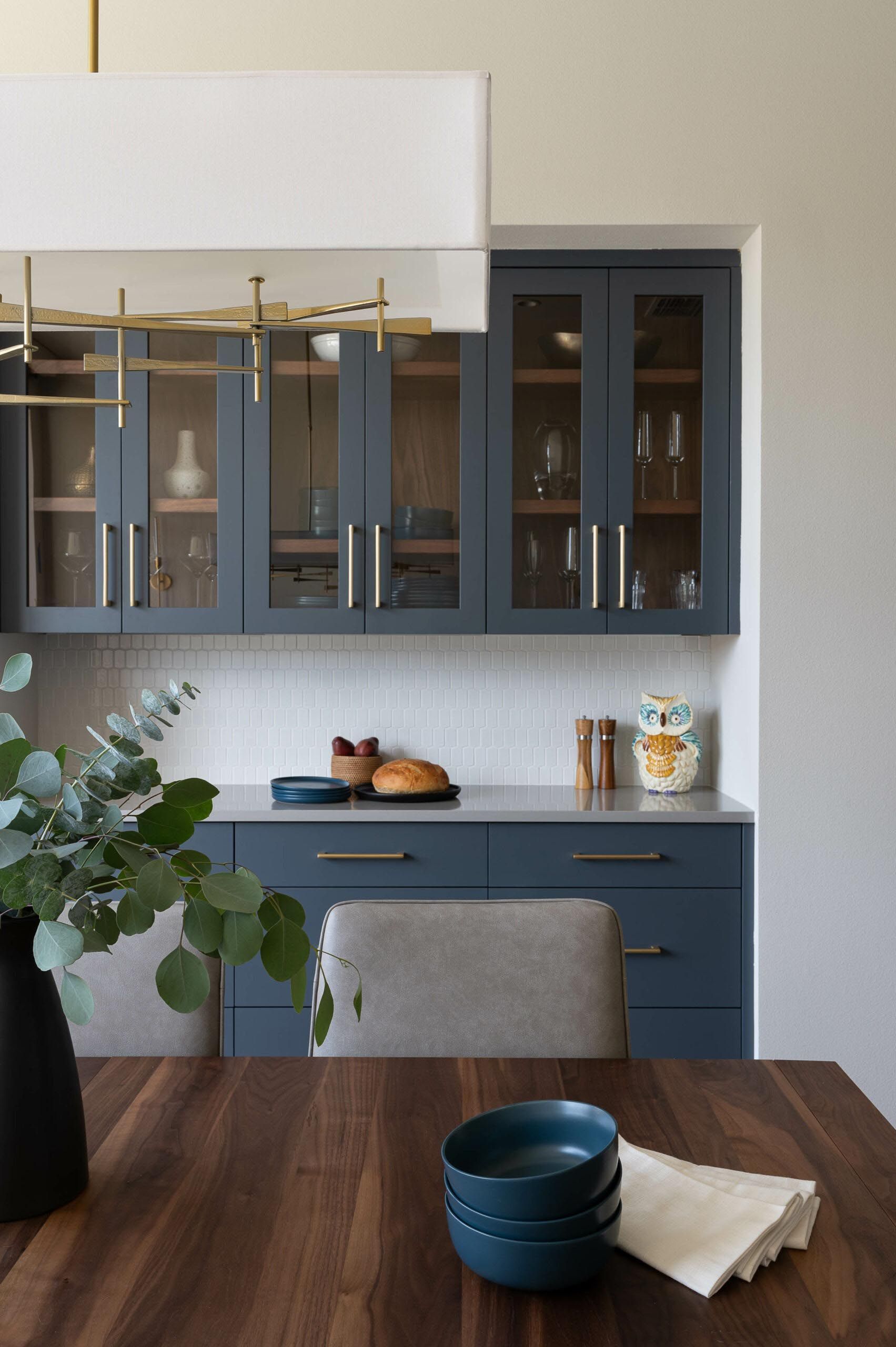 Blue kitchen cabinets with glass doors, white tile backsplash, wooden dining table, and green eucalyptus.