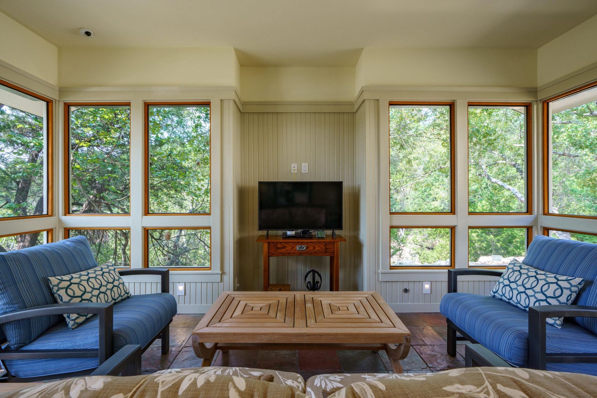 Living room with blue armchairs, wooden table, and TV surrounded by windows overlooking trees.