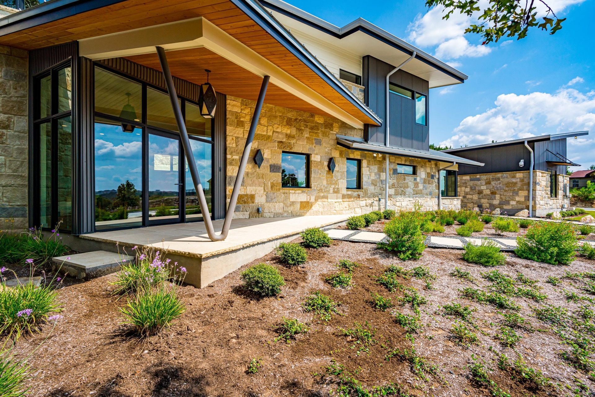 Modern house with stone and wood exterior, large windows, and landscaped yard under a blue sky.