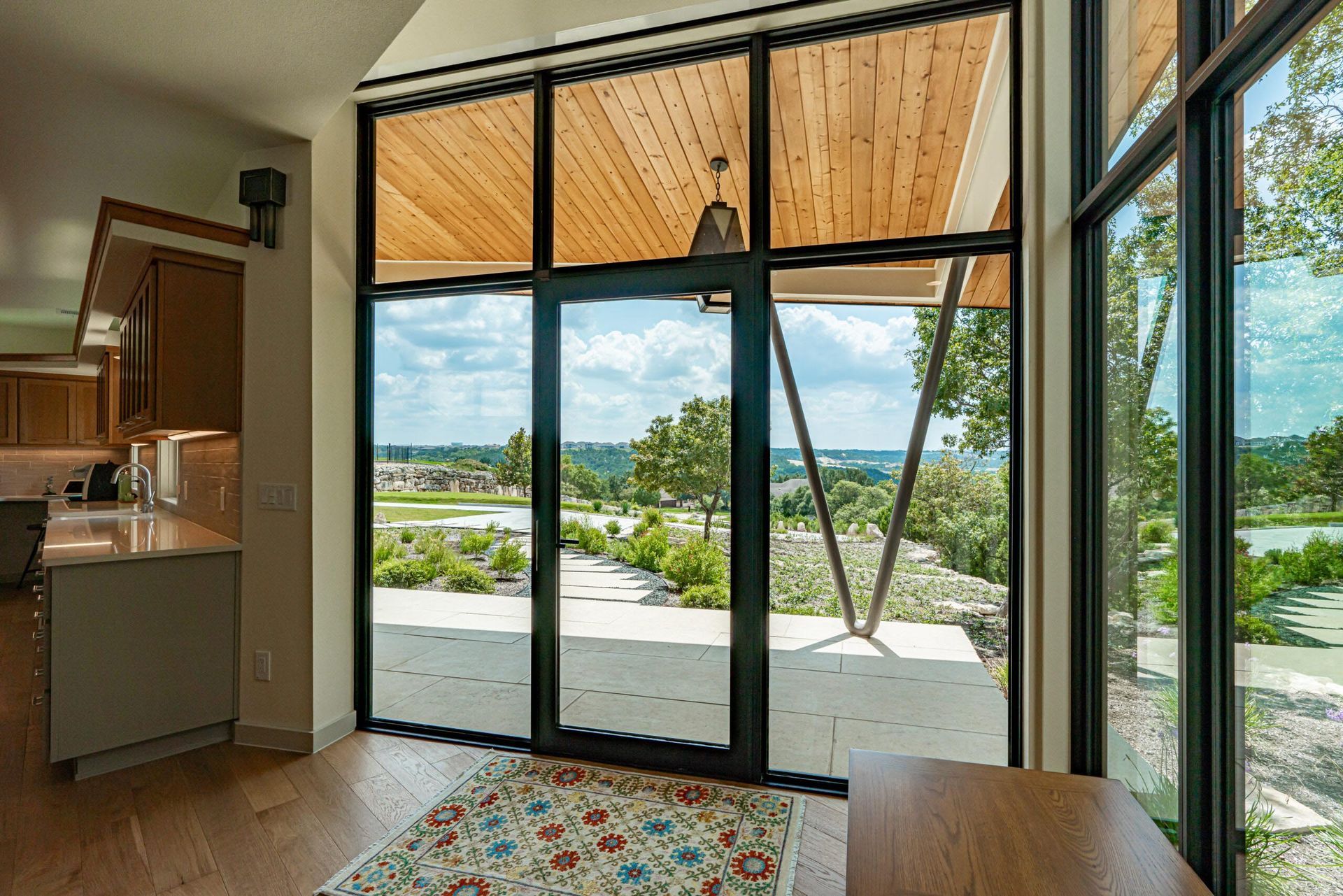 Interior view of modern home, large windows, scenic outdoor view. Wooden ceiling, gray kitchen, area rug.