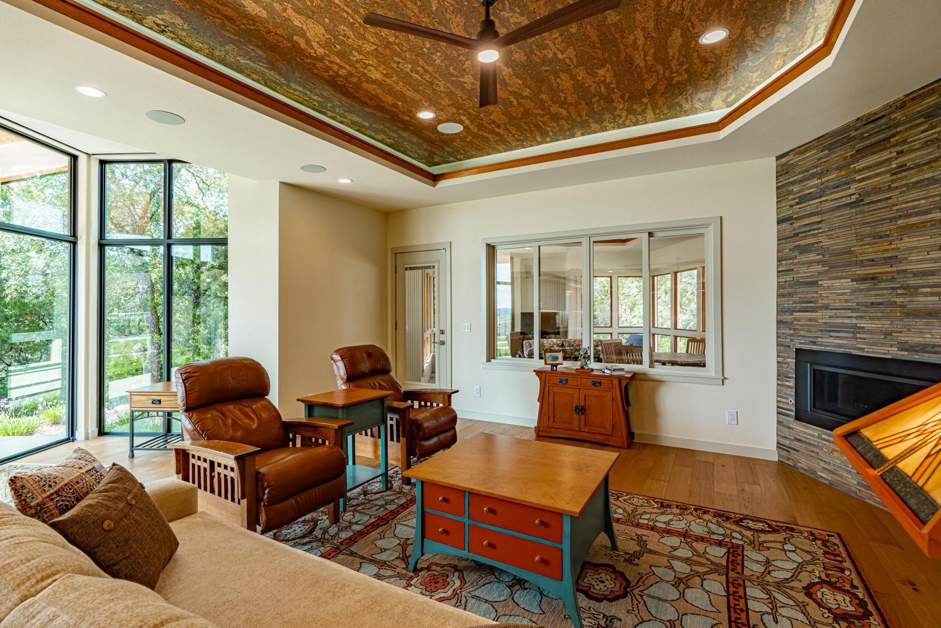 Living room with brown leather chairs, fireplace, patterned rug, and large windows overlooking greenery.